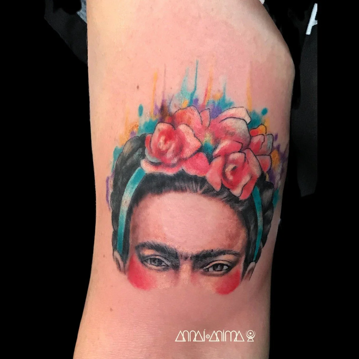 A colorful tattoo of Frida Kahlo's portrait with pink roses in her hair, vibrant splashes of watercolor around her head, and stylized symbols at the bottom.