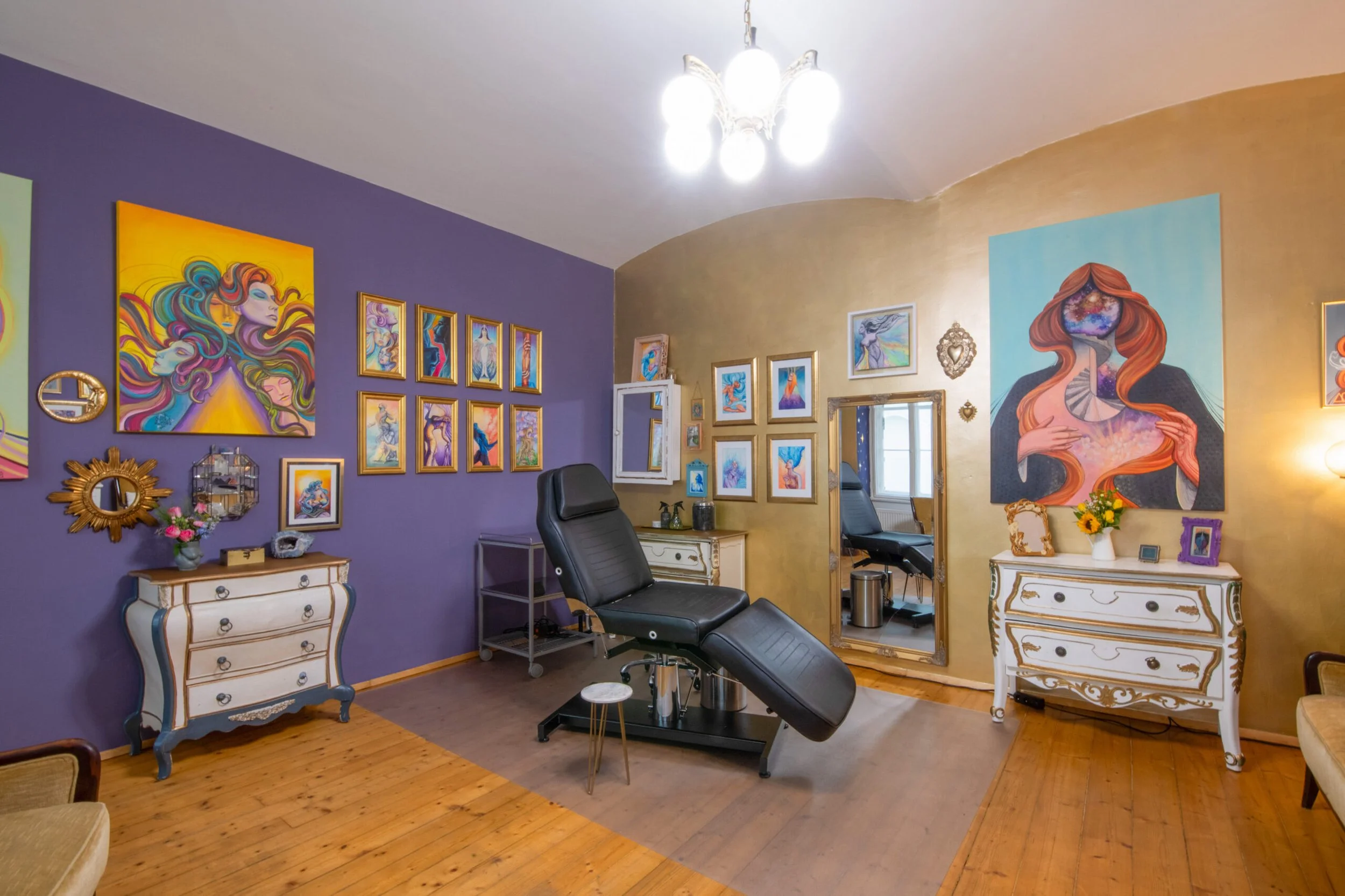A cozy tattoo studio with purple and beige walls decorated with colorful artwork. There is a black adjustable chair in the center, a large mirror, and ornate white dressers with flowers and picture frames. Tatuarium tattoo studio in Vienna, austria