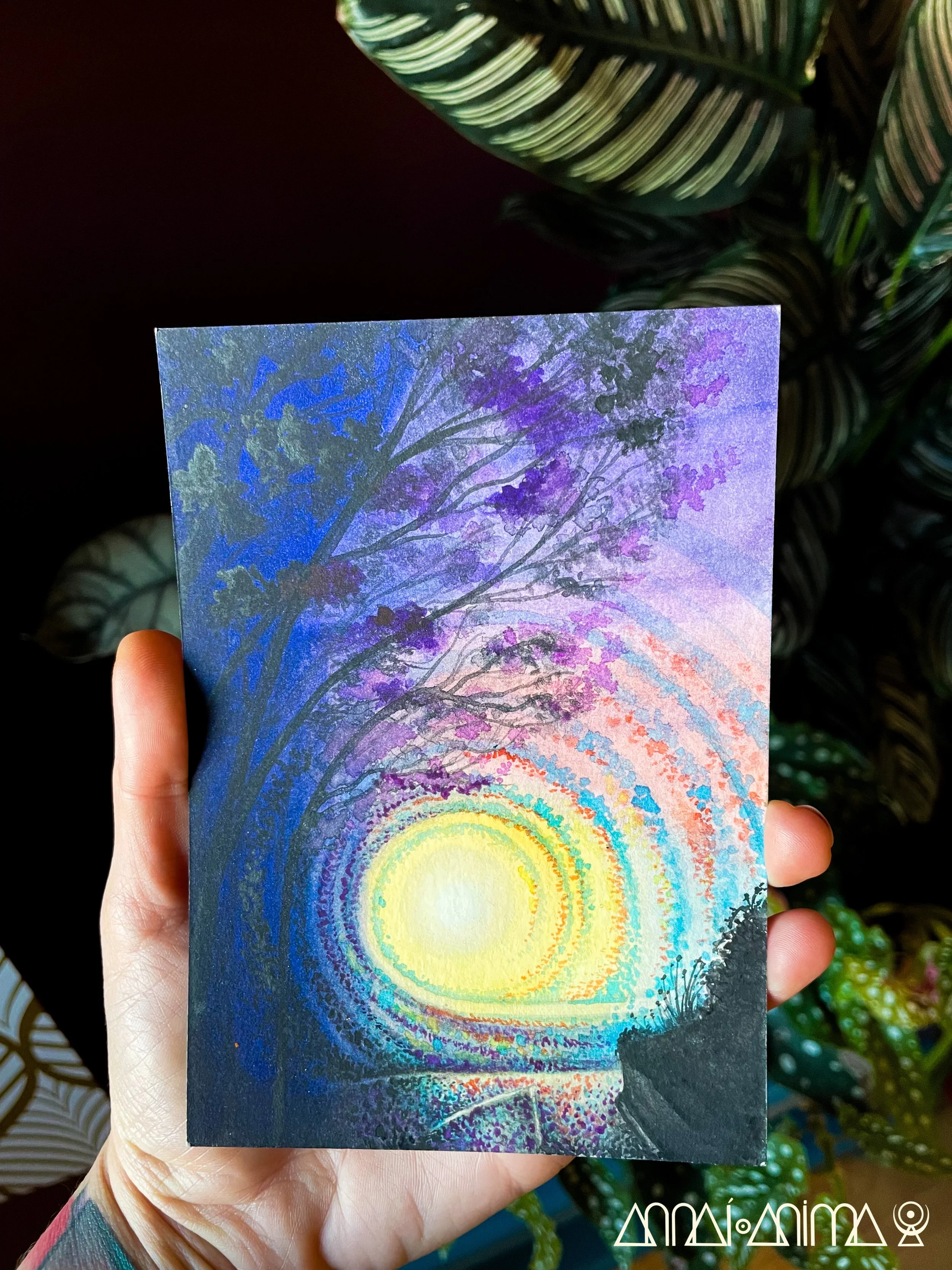 Hand holding a colorful painting of a sunset or sunrise with concentric circles of vibrant colors and purple trees in the foreground