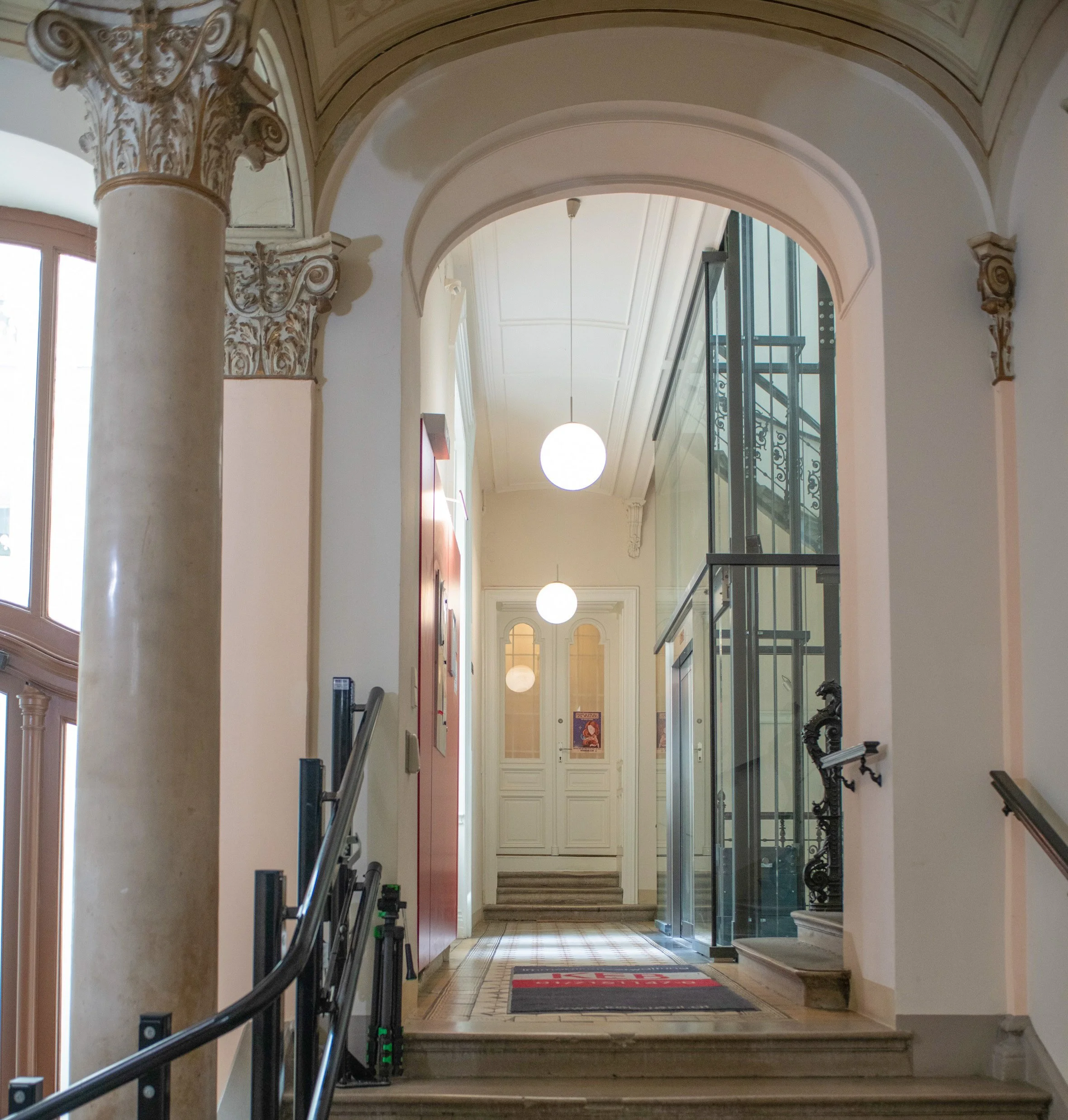 Interior view of a historic building with decorative columns, glass elevator, stairways, and white double doors at the end of a hallway with hanging round light fixtures. Tatuarium tattoo studio in Vienna, Austria