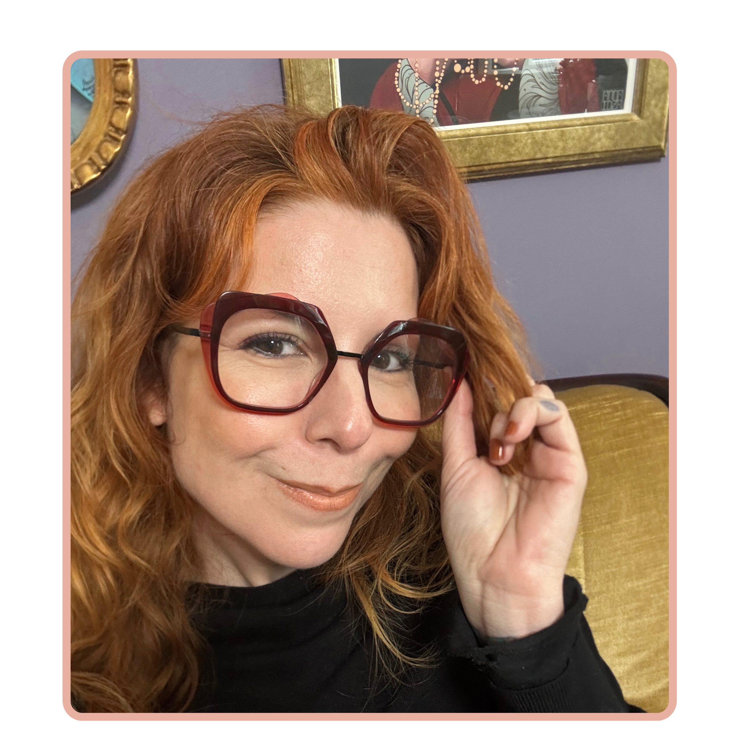 Annai Anima A woman with red hair and large red glasses smiling at the camera, sitting on a yellow couch with purple walls and framed artwork in the background.