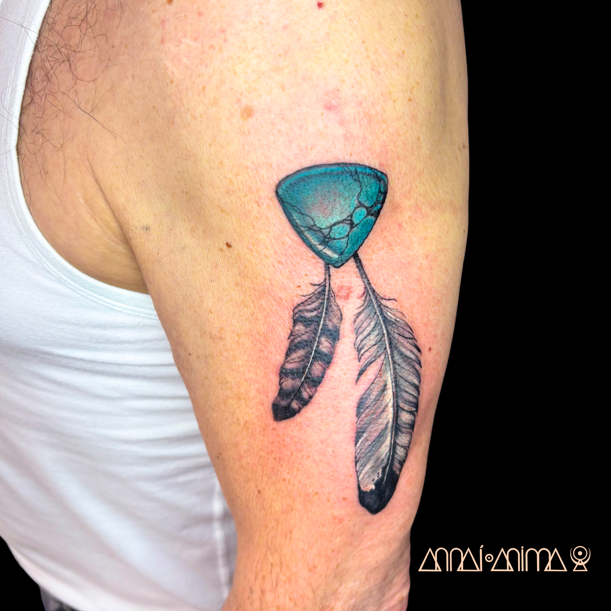 Tattoo of a blue and black stone and two feathers on a person's upper arm.
