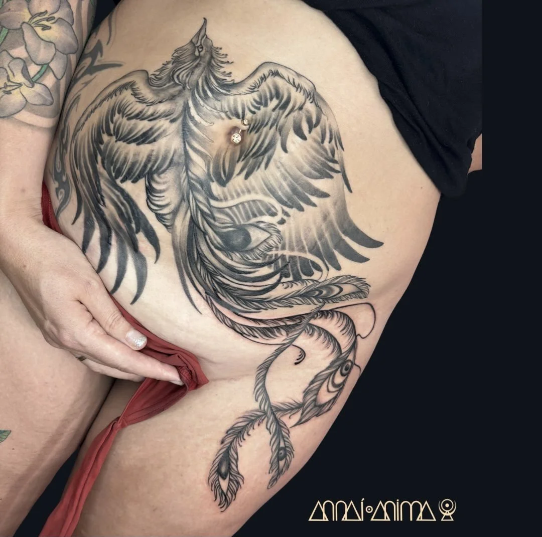 A large black and gray tattoo of a phoenix with detailed feathers covering the upper thigh and hip, with a person's hand pulling down a red garment near the tattoo.