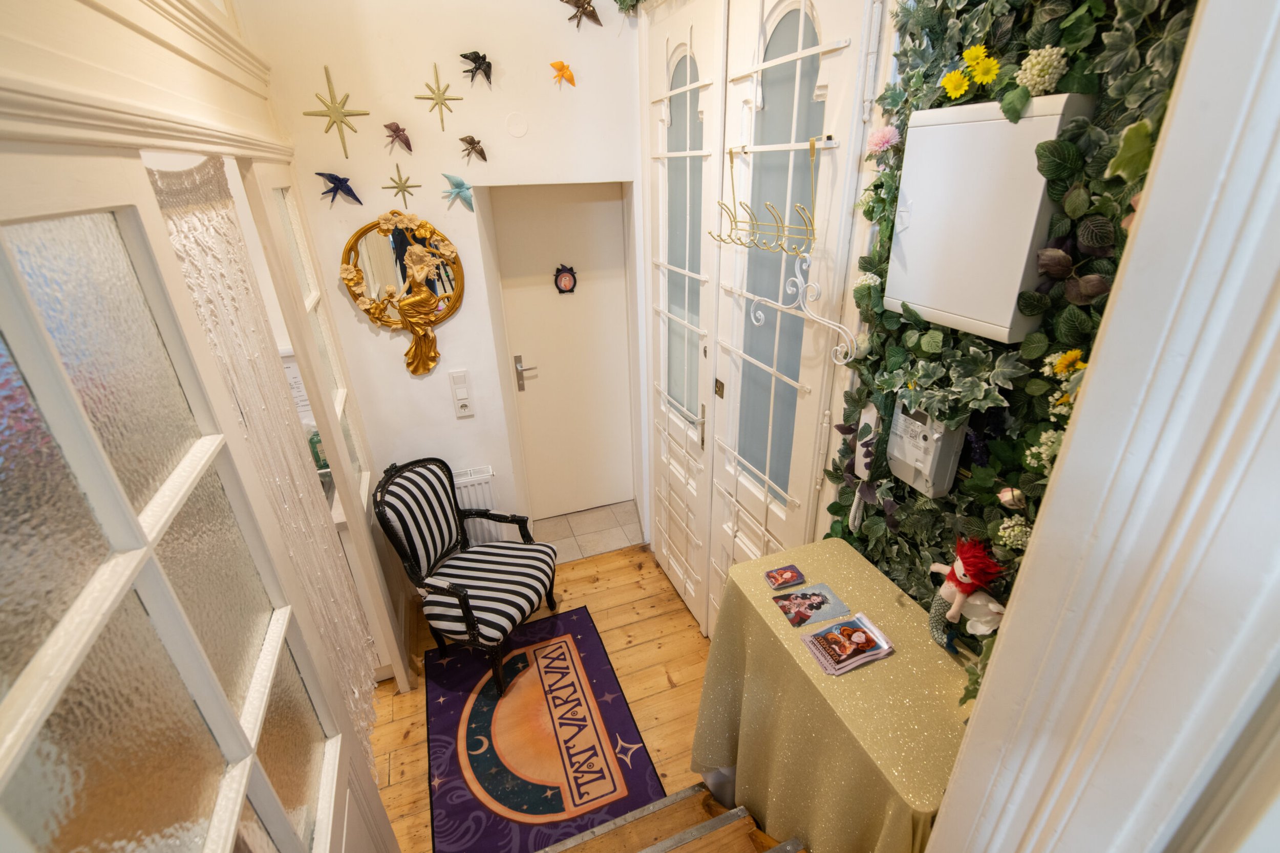 Decorative indoor entryway with a black and white striped chair, rainbow-colored zodiac rug, floral wall decor, mirror with gold adornment, and a table with photos, along with a door having glass panels. Tatuarium tattoo studio in Vienna, Austria