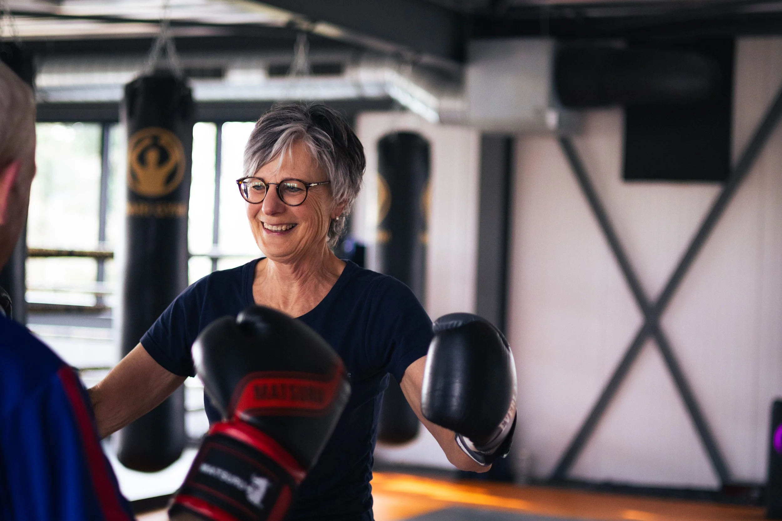 Vrouw in een sportschool met bokshandschoenen die aan het oefenen is, lachend en samen met een andere persoon.