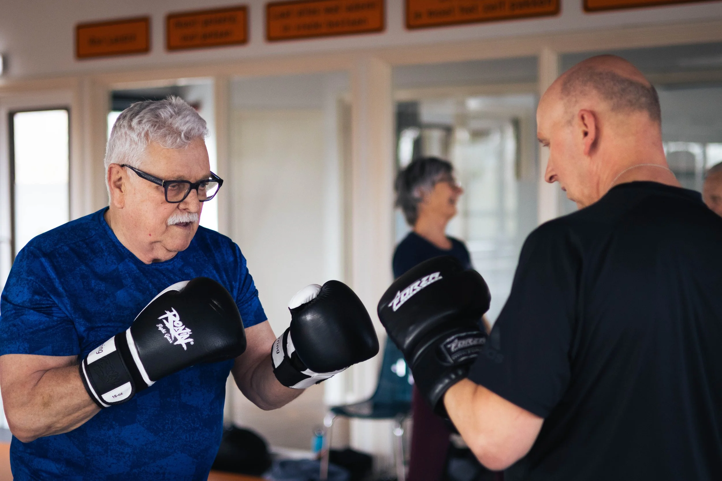 Twee oudere mannen in een boxing gym oefenen met bokshandschoenen, terwijl een vrouw in de achtergrond kijkt.