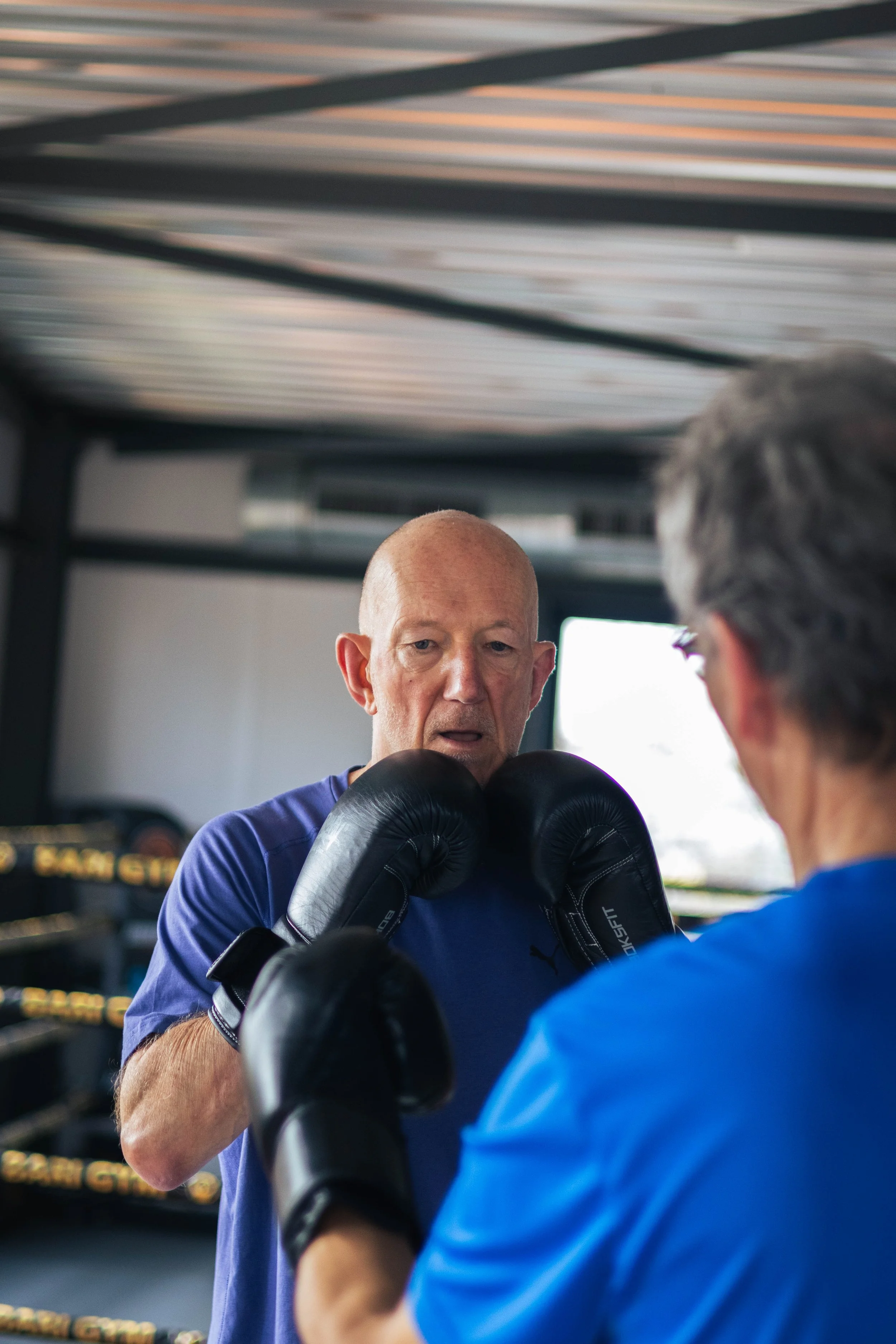 Twee mannen oefenen boksen in een sportschool. De man in de voorgrond draagt bokshandschoenen en een blauwe sportoutfit, terwijl de man in de achtergrond hem lijkt instructies te geven.