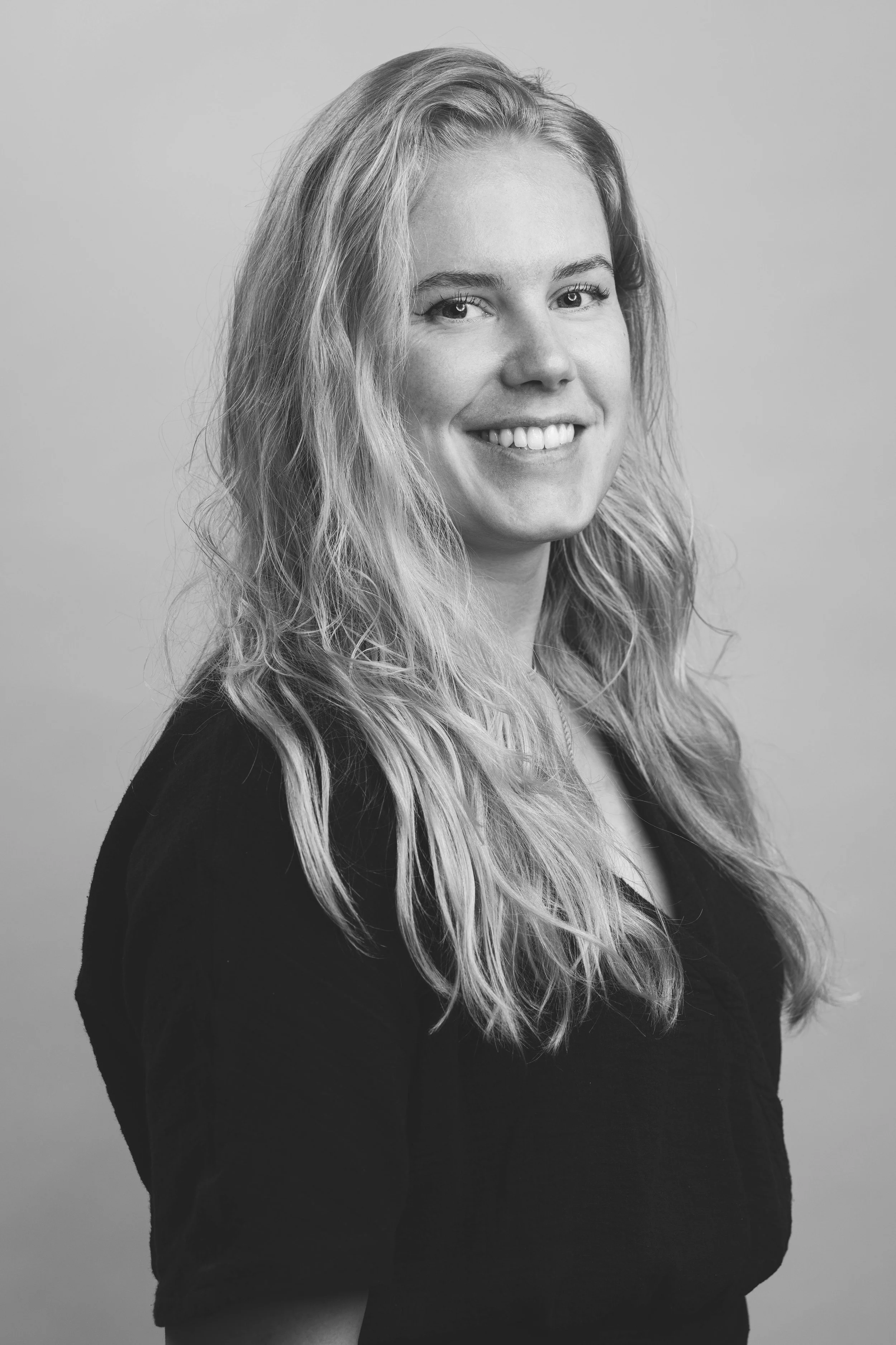 Zwart-wit portret van een lachende vrouw met lange, golvende haar, gekleed in een donkere top, tegen een effen achtergrond.