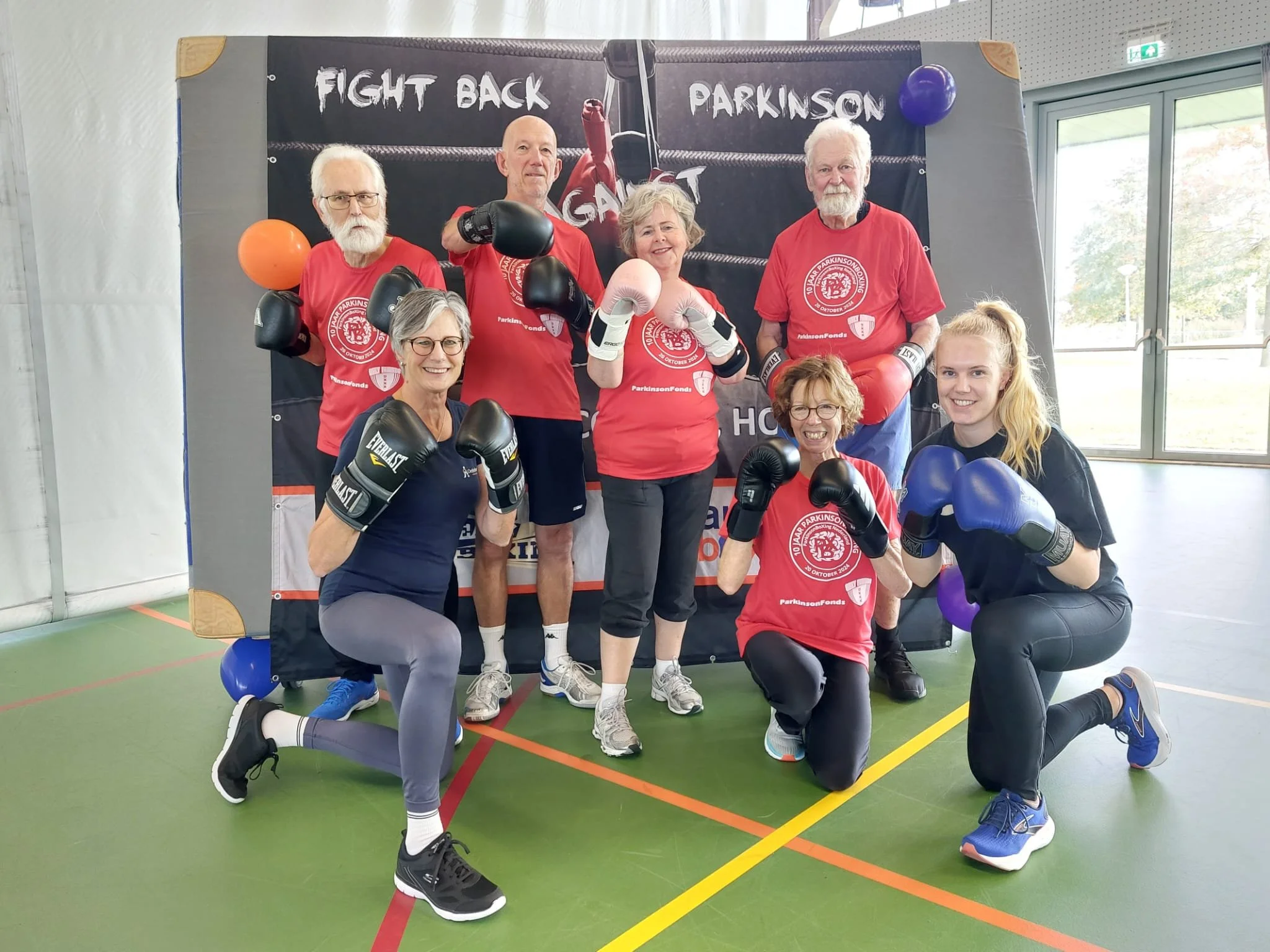 Een groep van negen mensen in een sportschool, gekleed in sportkleding en bokshandschoenen, poseert voor een achtergrond met tekst 'Fight Back Parkinson'.