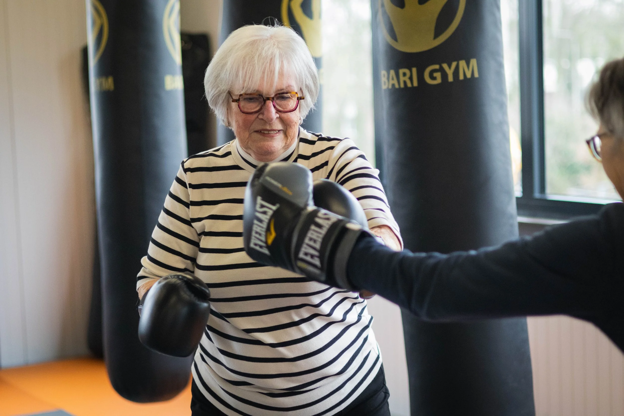 Een oudere vrouw met een zwart-wit gestreepte overhemd die in een sportschool traint met bokshandschoenen, terwijl een instructeur haar helpt.