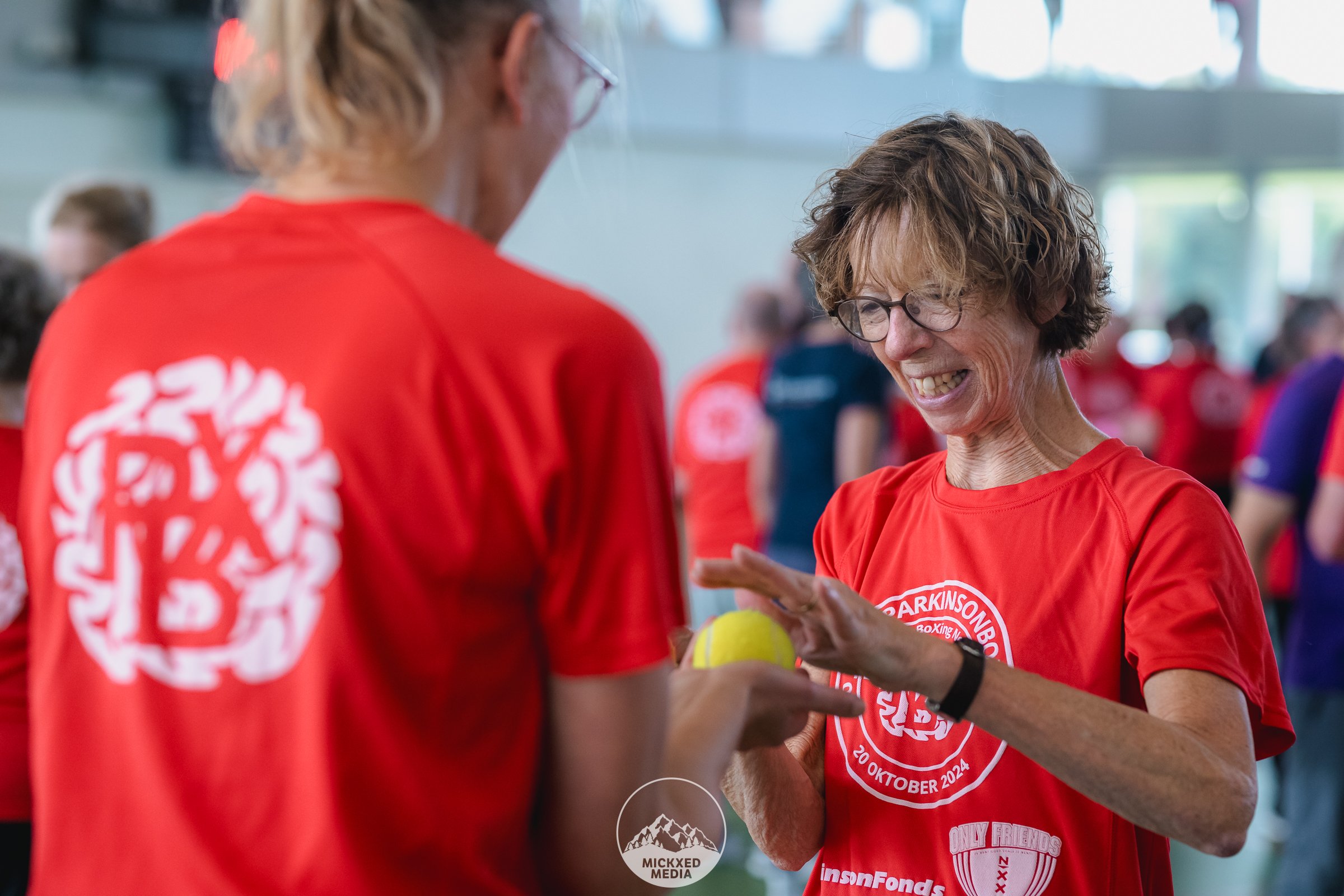 Twee mensen in rood sportkleding die met elkaar praten en lachen, in een grote binnenhal met andere mensen op de achtergrond.