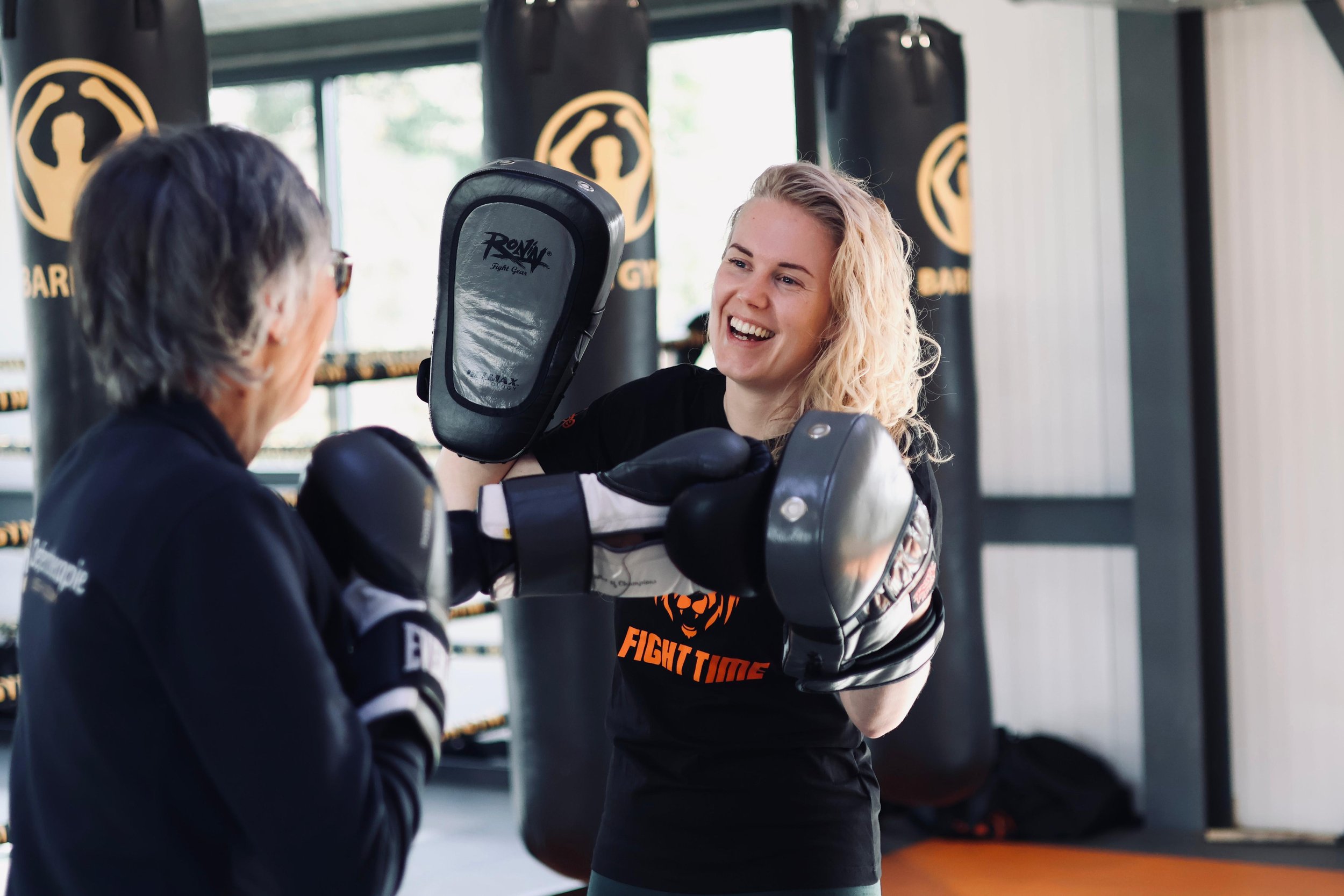 Vrouw die in een vechtsportclub met een treinster in een bokszak traint, lachend en met bokshandschoenen aan.