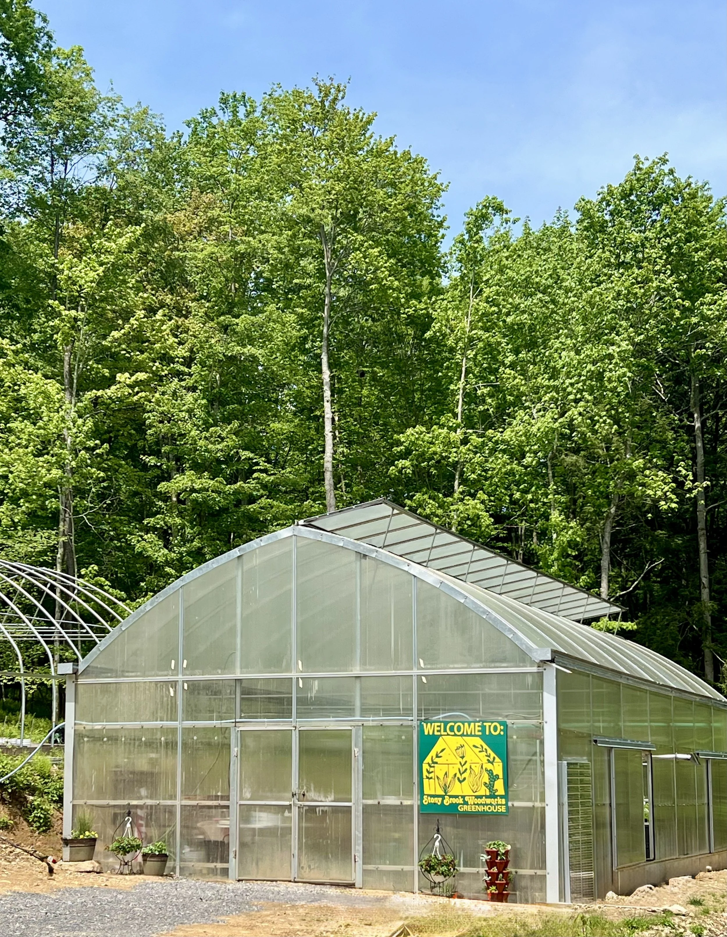 A greenhouse with a sign that reads "Welcome to Stony Brook Woodworks Greenhouse" is located in front of lush green trees on a sunny day.