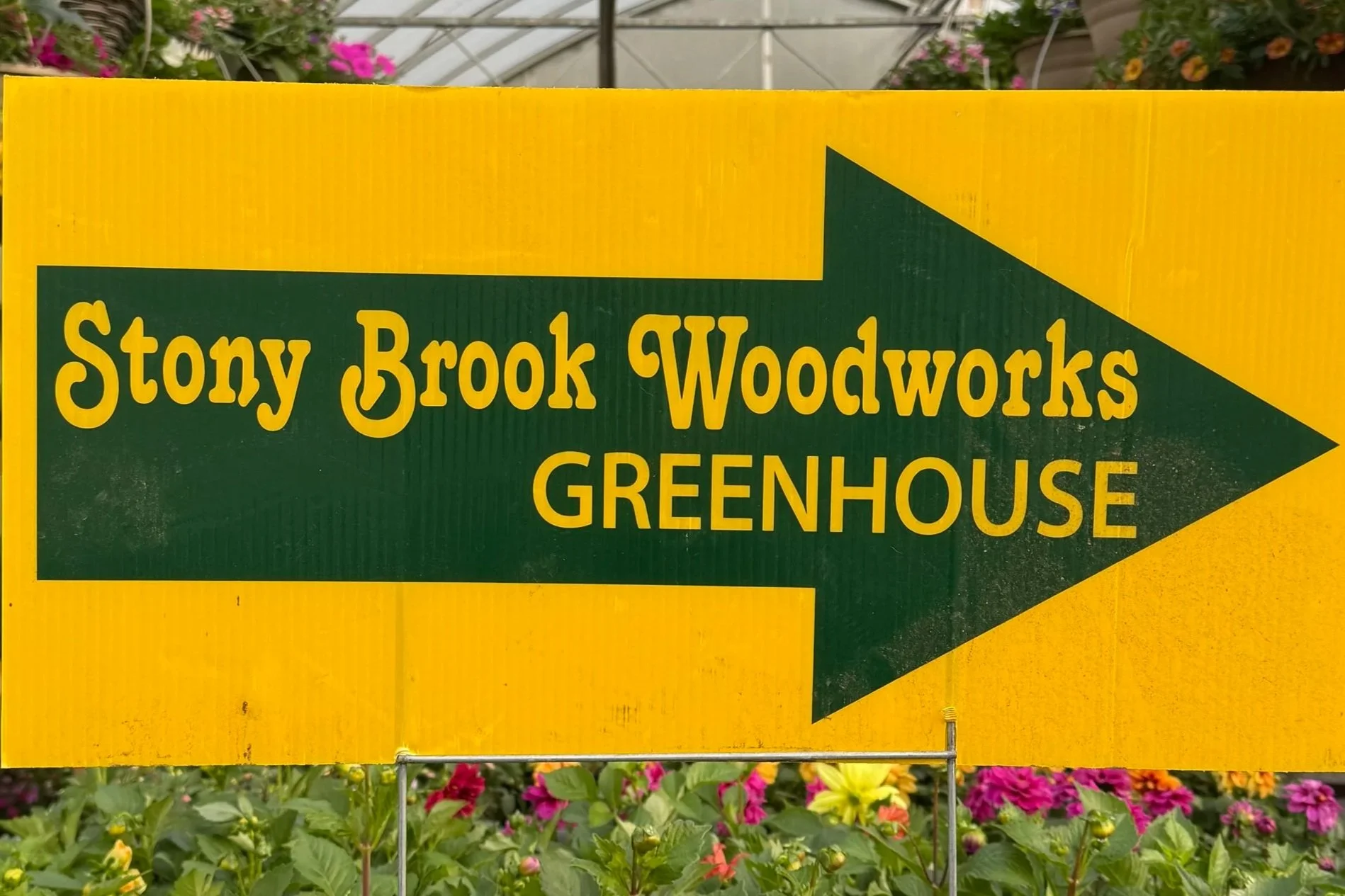 Yellow sign with green arrow pointing right for Stony Brook Woodworks greenhouse, surrounded by colorful flowers.