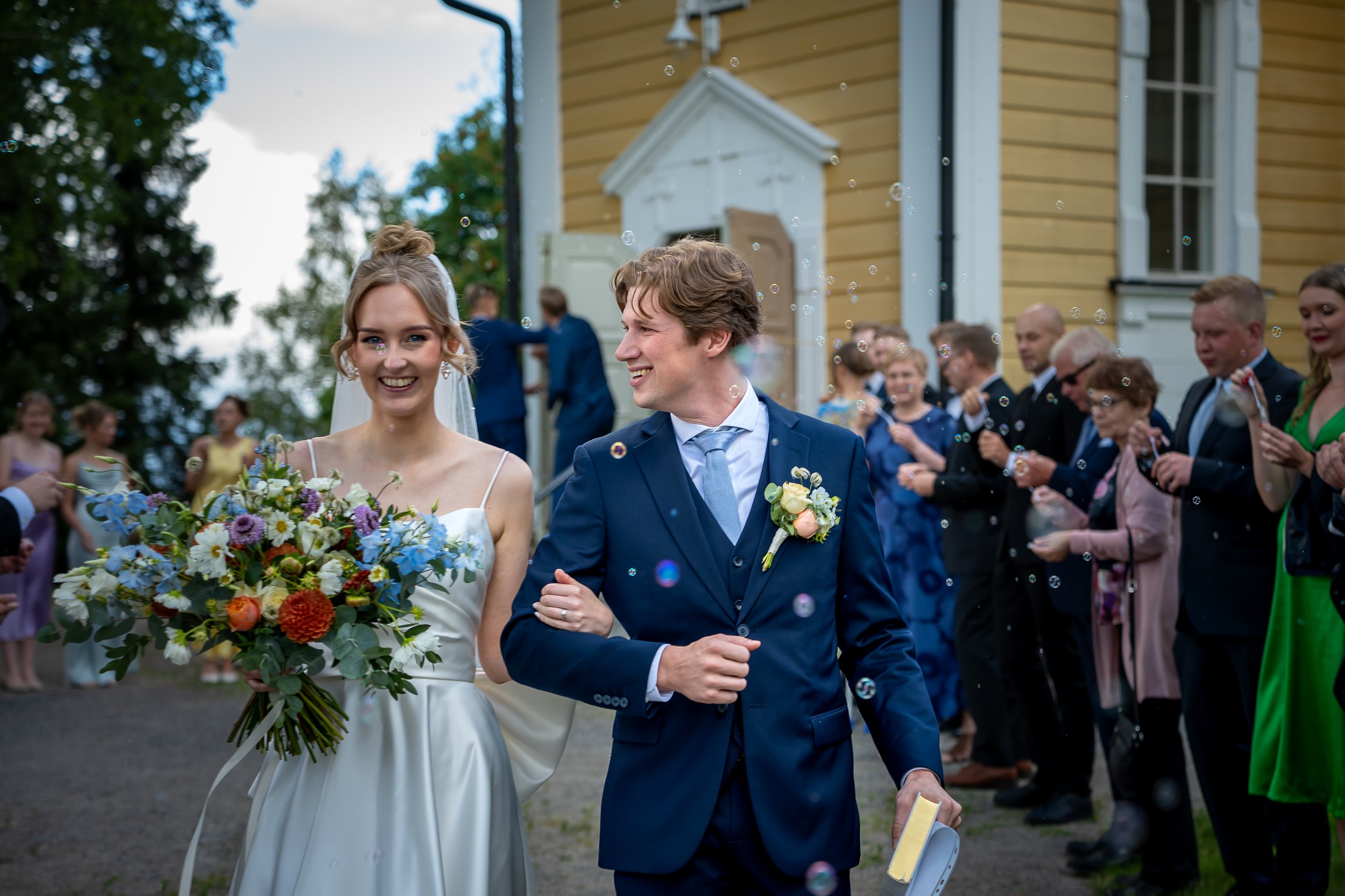 Hääpari kävelee yhdessä käsikynkässä hääjuhlassa, jonka osallistujat katsovat ja ihmettelevät. Taustalle näkyy vanha puutalo ja populaa juhlasta.