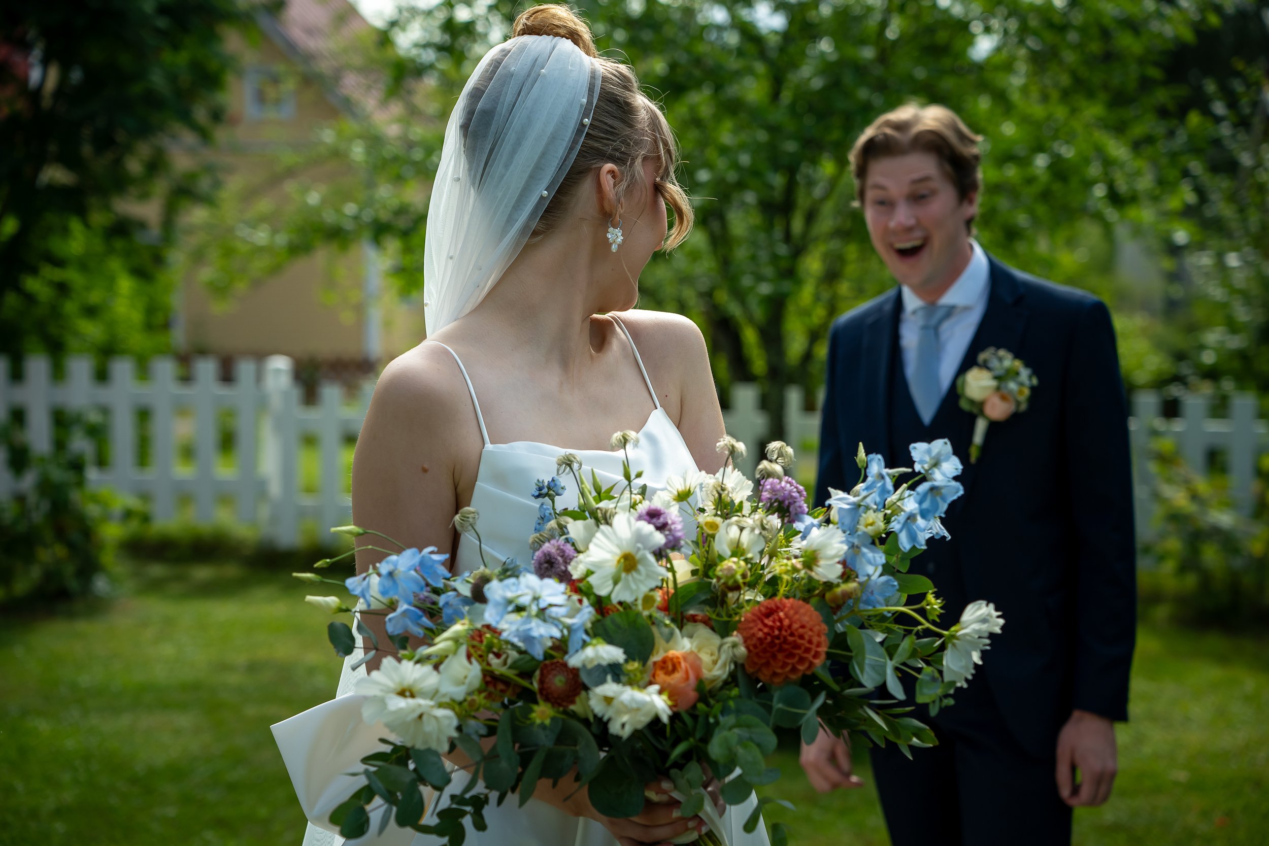 Hääpari iloitsee toisistaan puutarhassa, nainen pidättää kukkakimppua ja mies nauraa.