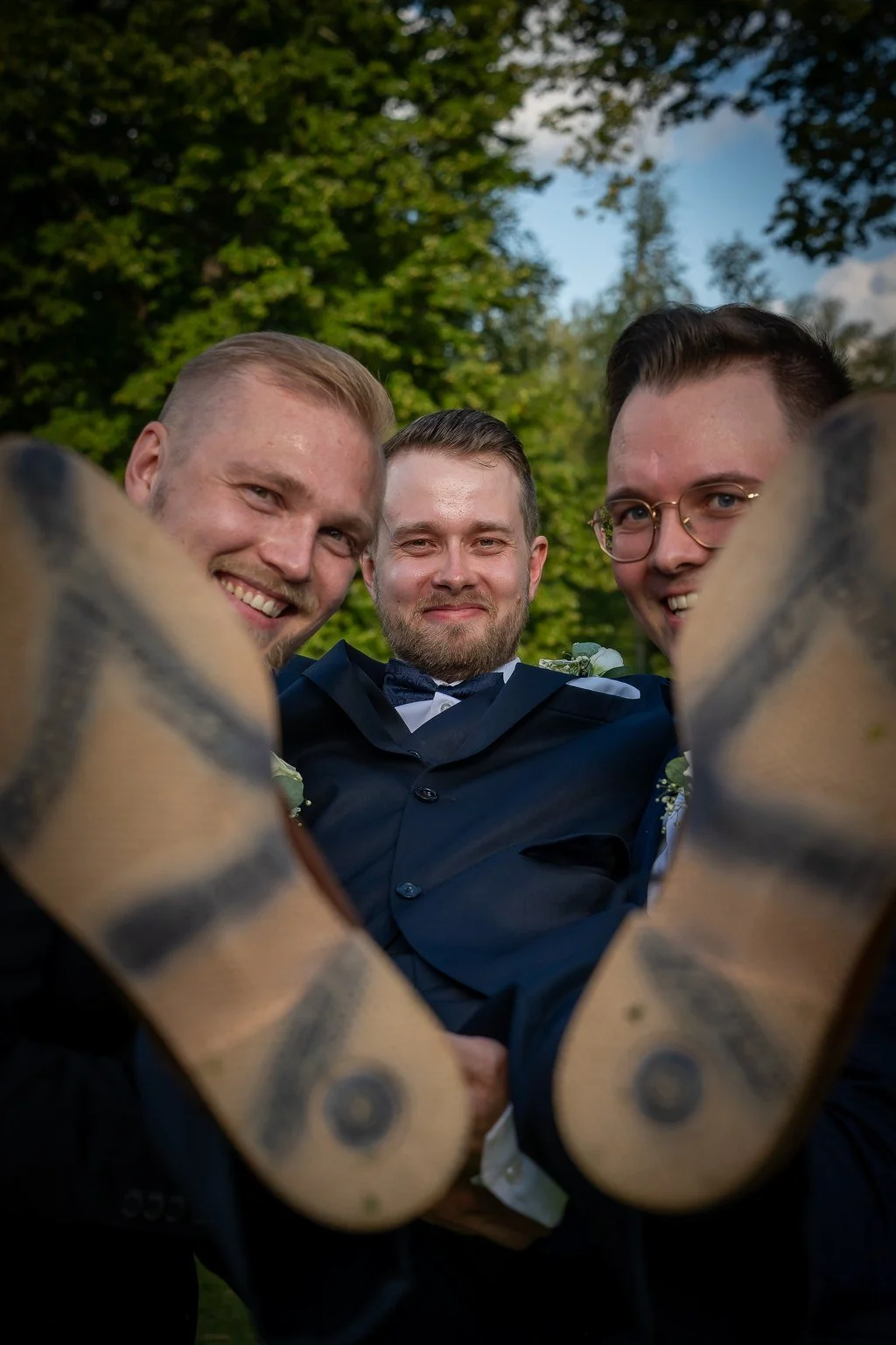 Kolme miestä hymyilevät läheltä katsottuna ulkona, taustalla puita ja sininen taivas.