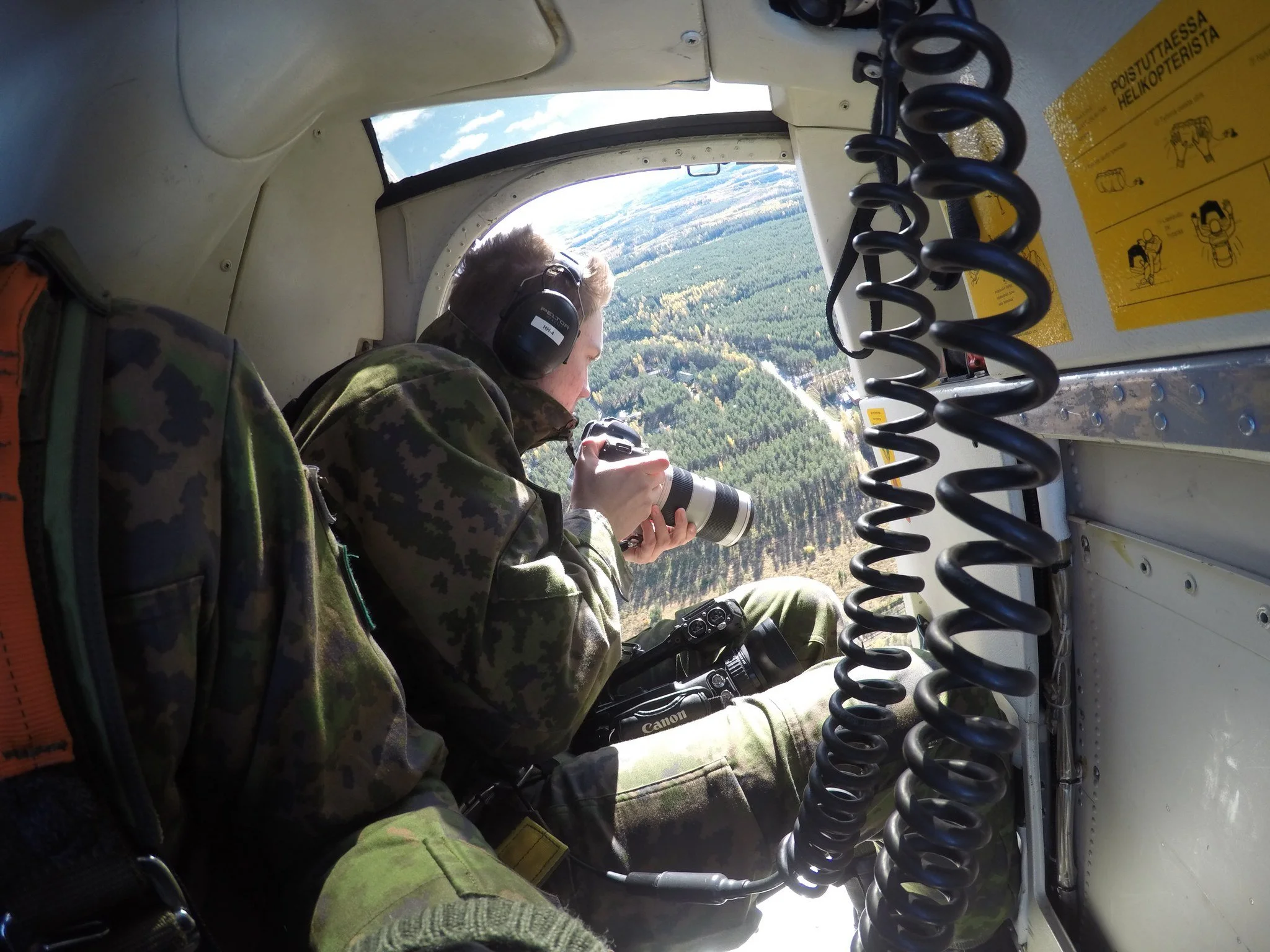 Henkilö kuvattuna lentokoneen sisällä, katsellen ulos ja ottamassa valokuvia, ympärillään lentokoneseiniä ja kuulokkeet.