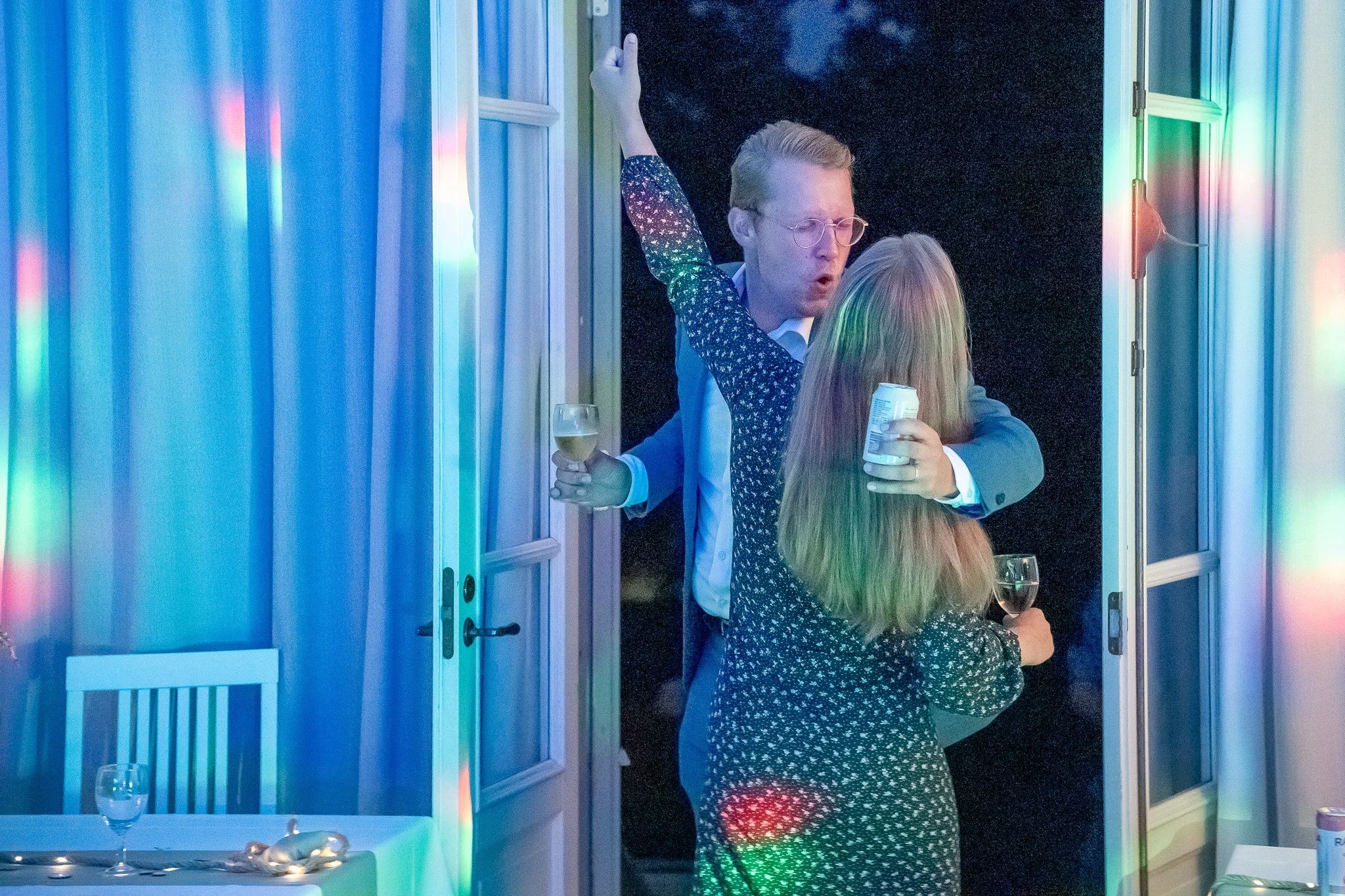 Kaksi ihmistä halaavat toisiaan juhlassa, toinen pitää lasillista juotavaa ja toinen tölkkiä, taustalla on avattu ovi ja ilmainen valolaite.