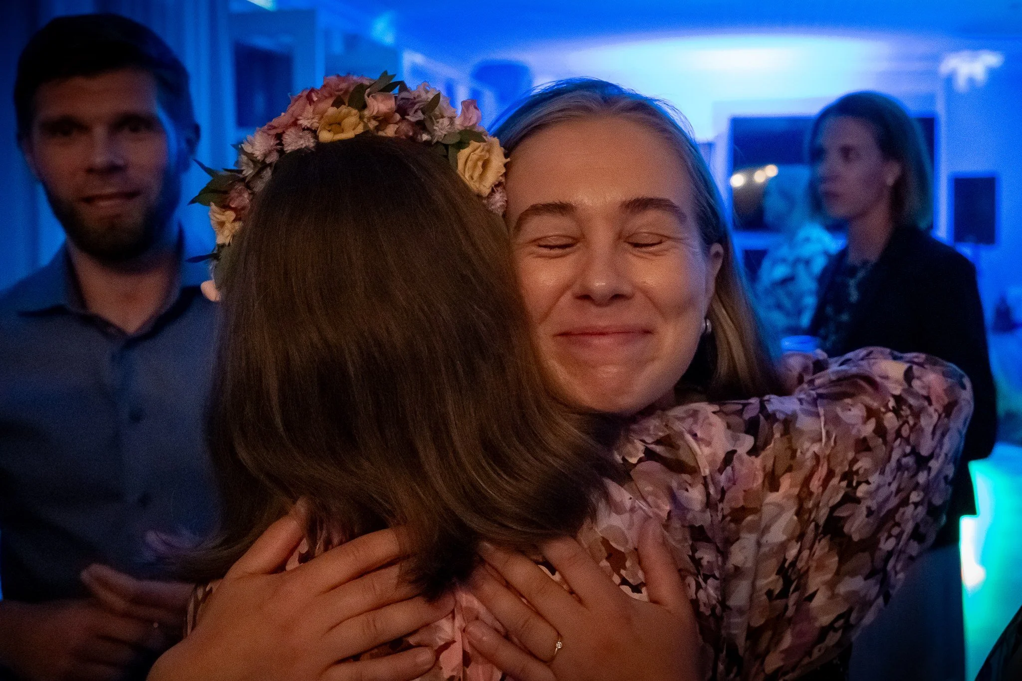 Kaksi naista halaavat iloisesti juhlatilassa, toinen naisista on iloisesti hymyilevä ja toisella on päähänsä kukkainen seppele, taustalla kolme ihmistä katsovat.