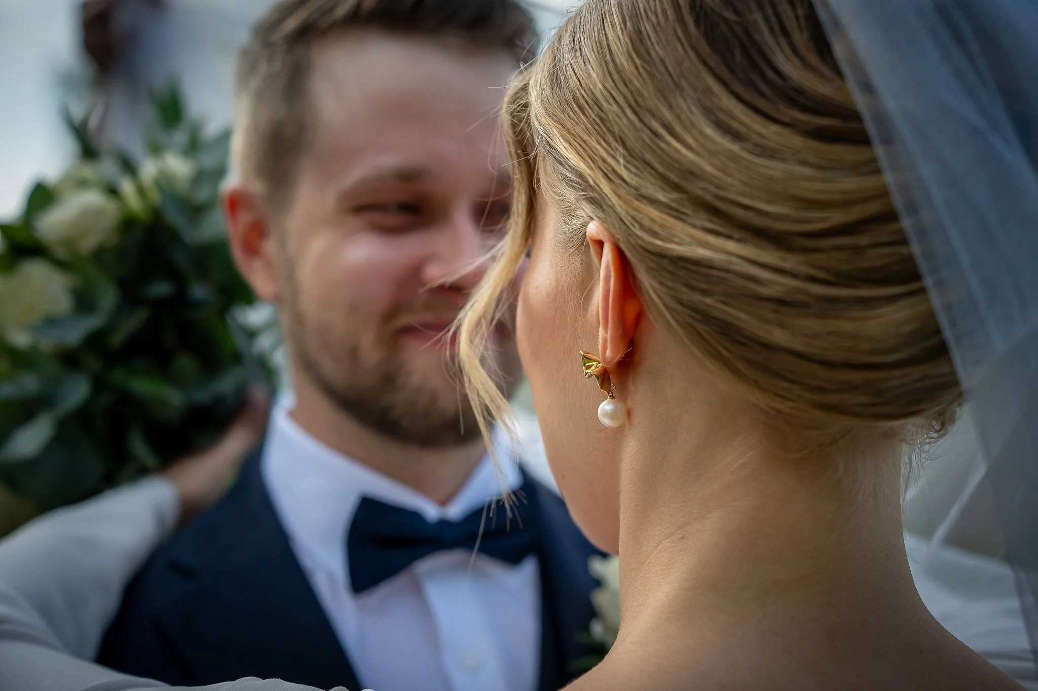 Hääpari katsoo toisiaan läheltä, taustalla vihreitä kasveja ja valkoisia kukkia.