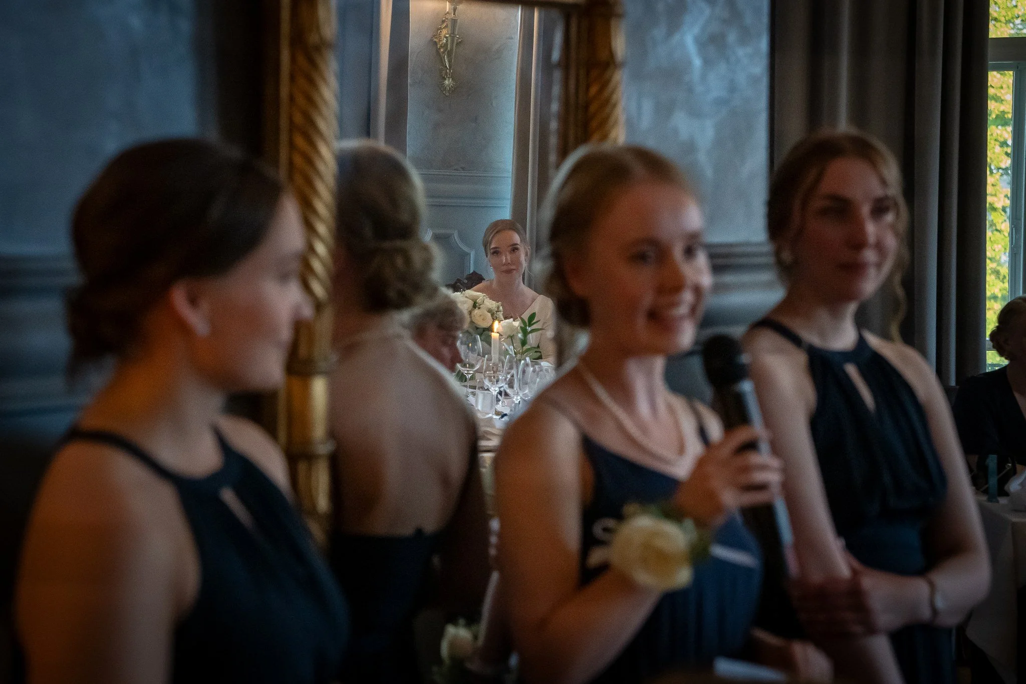 Koristelevi joukko naisia hameissa nanomaisemiin, taustalla näkyy nainen istumassa pöydässä, jossa on kukkia ja kynttilöitä, ja taustalla näkyy iso ikkuna ja kynttilä.