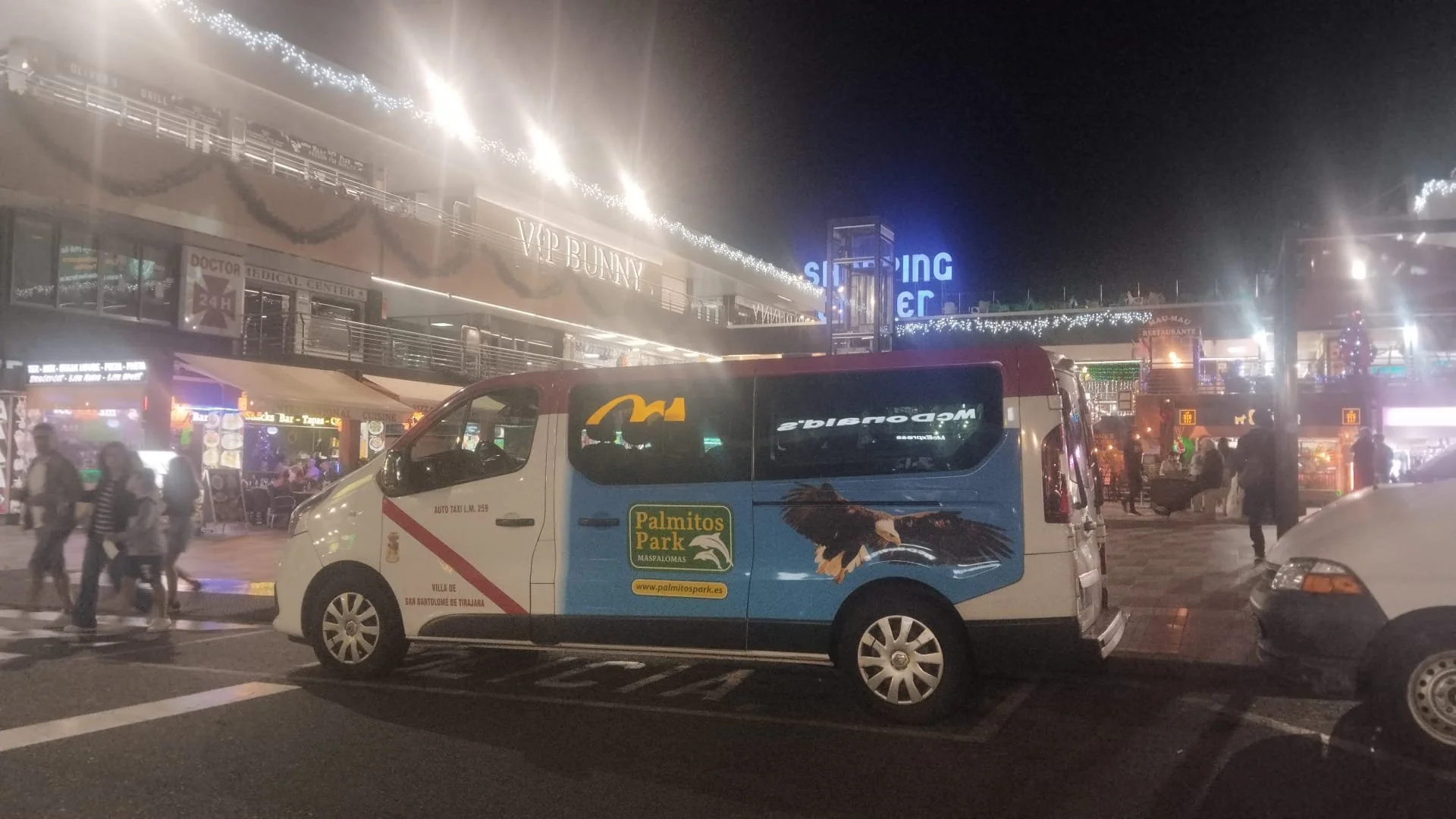 Imagen de una calle por la noche con un vehículo de transporte, un parque temático llamado Palmitos Park, tiendas y gente caminando.