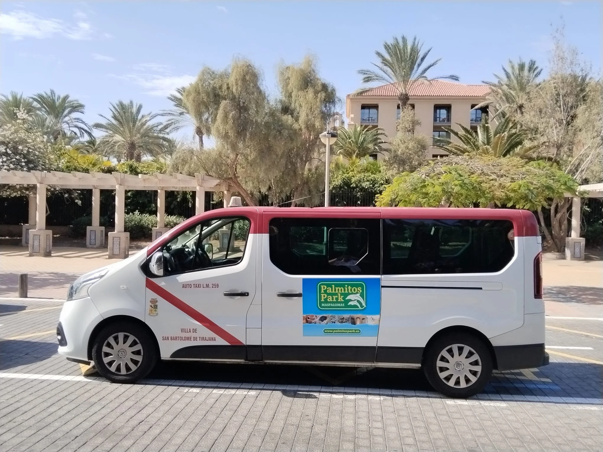 Vehículo de alquiler de color blanco con partes superiores en color rojo, con anuncios de Palmitos Park en la puerta y en la parte trasera, estacionado en un parque con árboles y un edificio de varias plantas en el fondo.