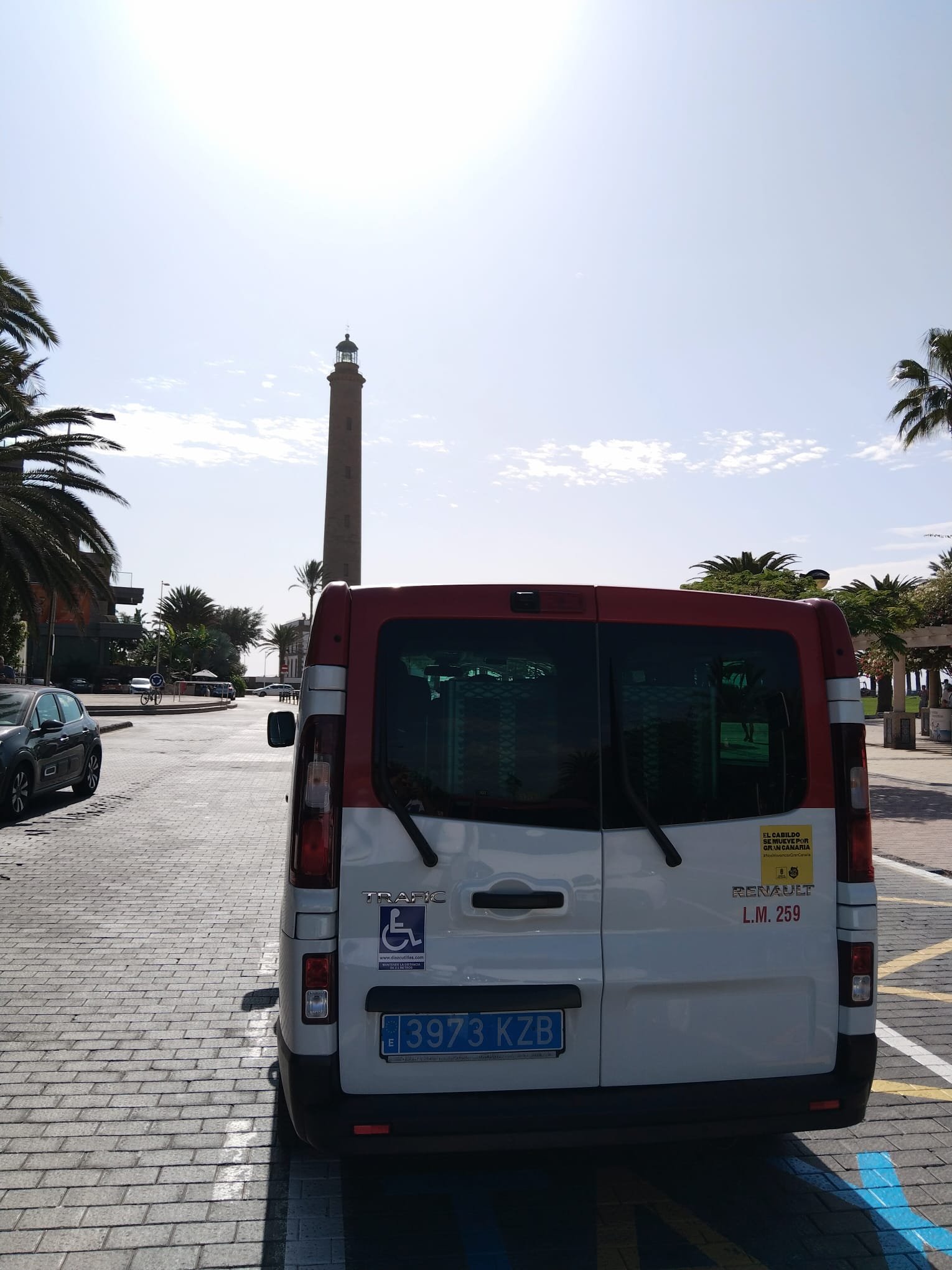 Estacionamiento con un vehículo de discapacitados, un faro y palmeras en un día soleado.