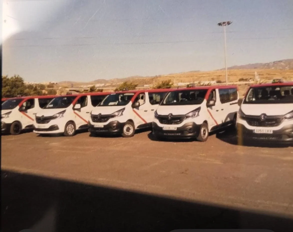 Varias furgonetas blancas con detalles rojos en una fila en un estacionamiento al aire libre, con un fondo de montañas y cielo despejado.