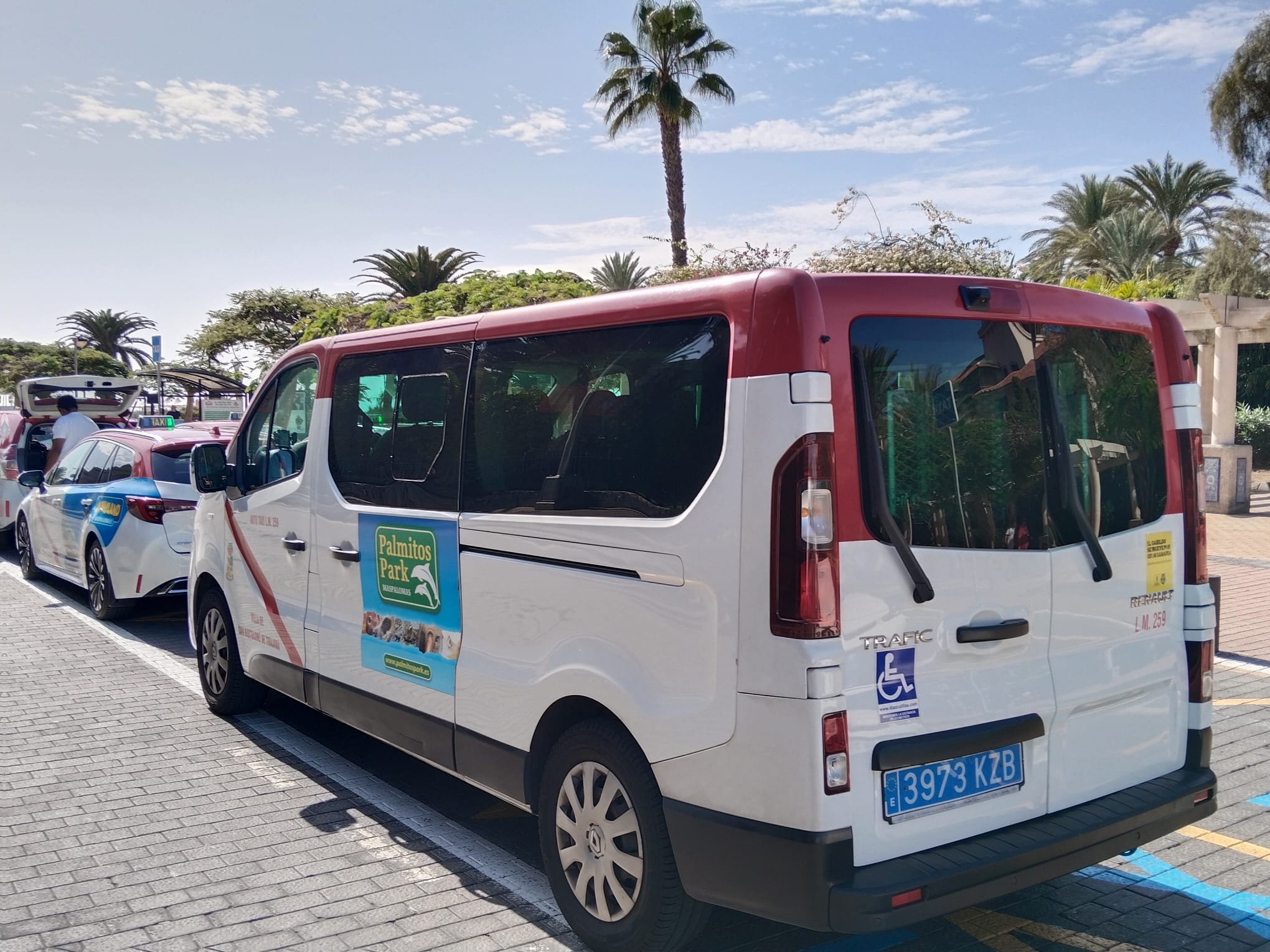 Furgoneta blanca y roja de transporte en un estacionamiento, con vigía de accesibilidad para discapacitados y cartel de anuncios turísticos.