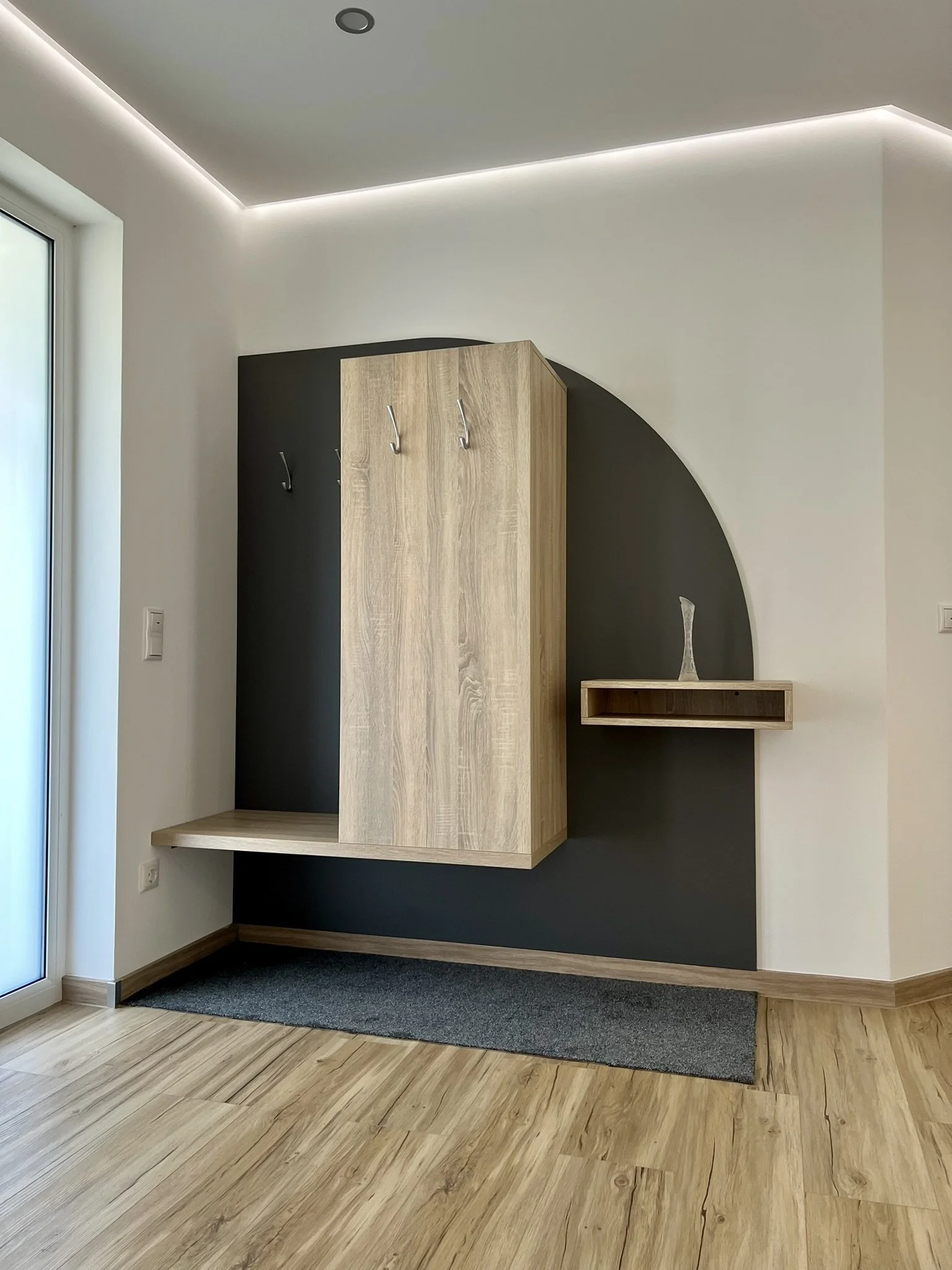 Moderner Eingangsbereich mit Holzfläche, Garderobe mit Haken, kleiner Wandnische mit Deko-Objekt, dunkler Teppich auf Holzboden, weiße Wände mit schwarzem Akzent, große Fenster