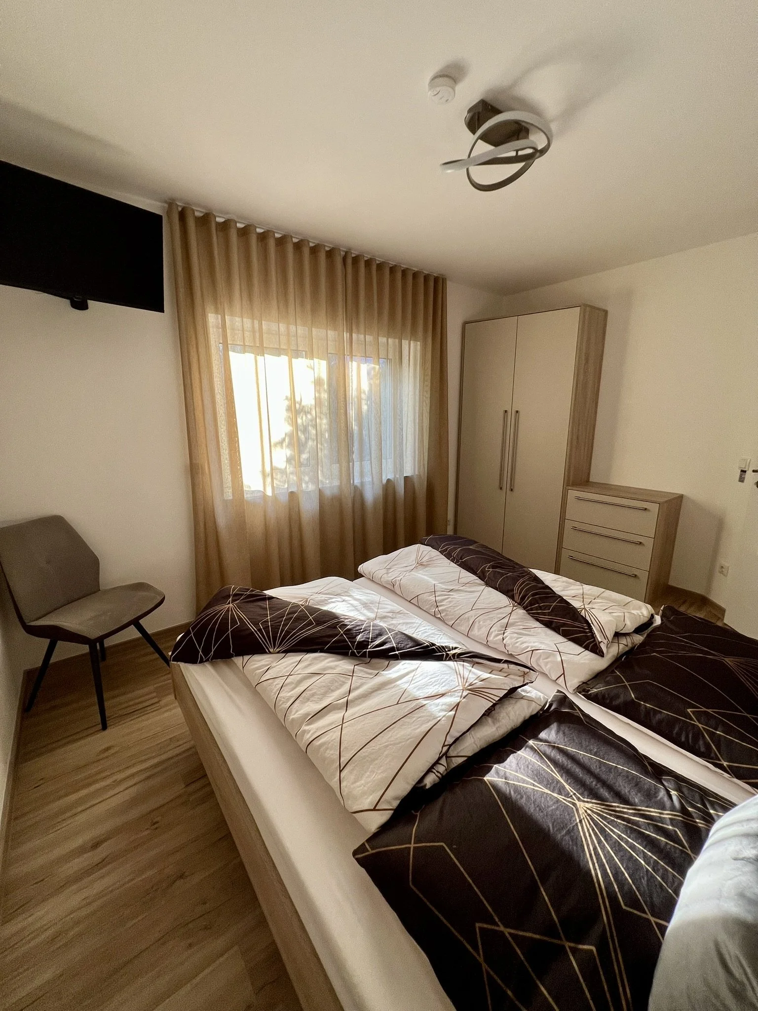 Schlafzimmer mit Bett, Kissen, Bettdecke, Nachttisch, Schrank, Fernseher und Stuhl. Vorhänge vor einem Fenster, Holzfußboden, weiße Wände.