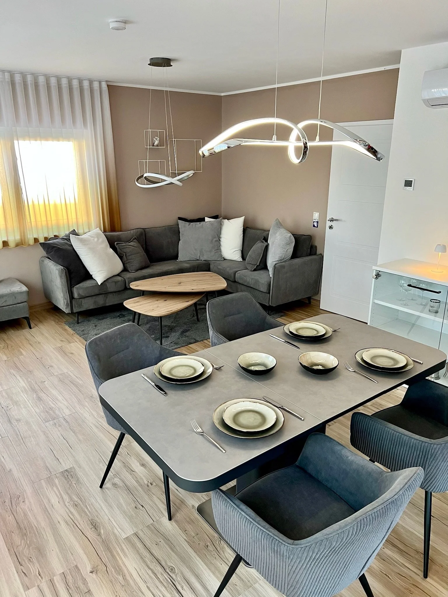 Ein Esstisch mit sechs Tellern und Besteck, umgeben von sechs Stühlen, im Vordergrund ein grauer Sessel, im Hintergrund ein Wohnzimmer mit Couch und Kaffeetisch, eine Lampe hängt von der Decke, ein Fenster mit gelben Vorhängen ist sichtbar.