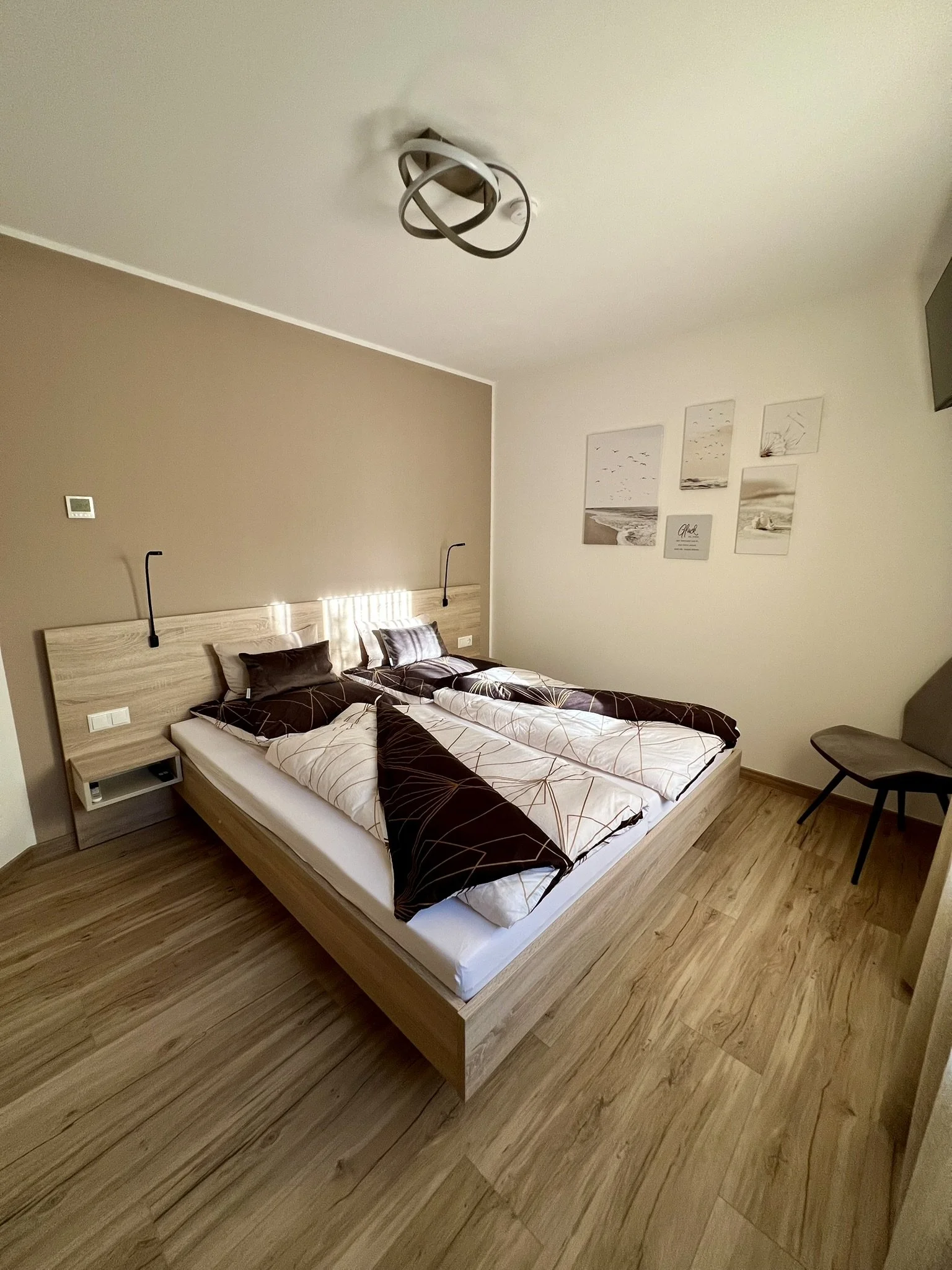 Ein Schlafzimmer mit einem Doppelbett, dunklen Bettwäsche, Holzfußboden, Wandbilder mit Strand- und Meeresmotiven, und einer schwarzen Stuhl in der Ecke.