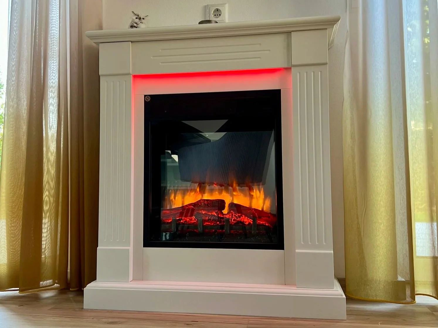 Elektrischer Kamin mit Flammen- und Glut-Design, in einem weißen Gehäuse, neben gelben Vorhängen.
