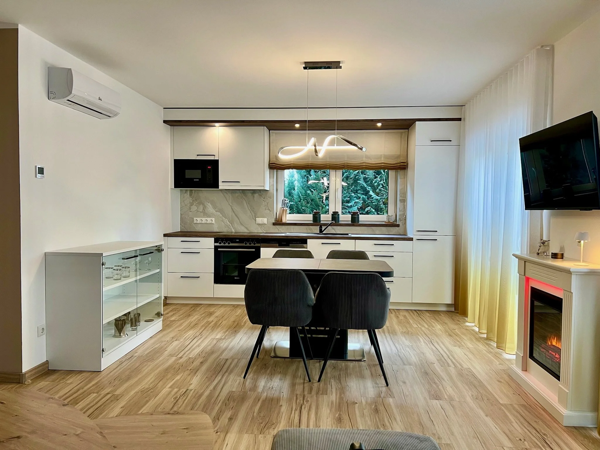 Modernes Wohnzimmer mit offener Küche, gemütlichem Essbereich und großen Fenstern mit Blick auf einen grünen Garten, Holzboden, Flachbildfernseher und elektrischem Kamin.