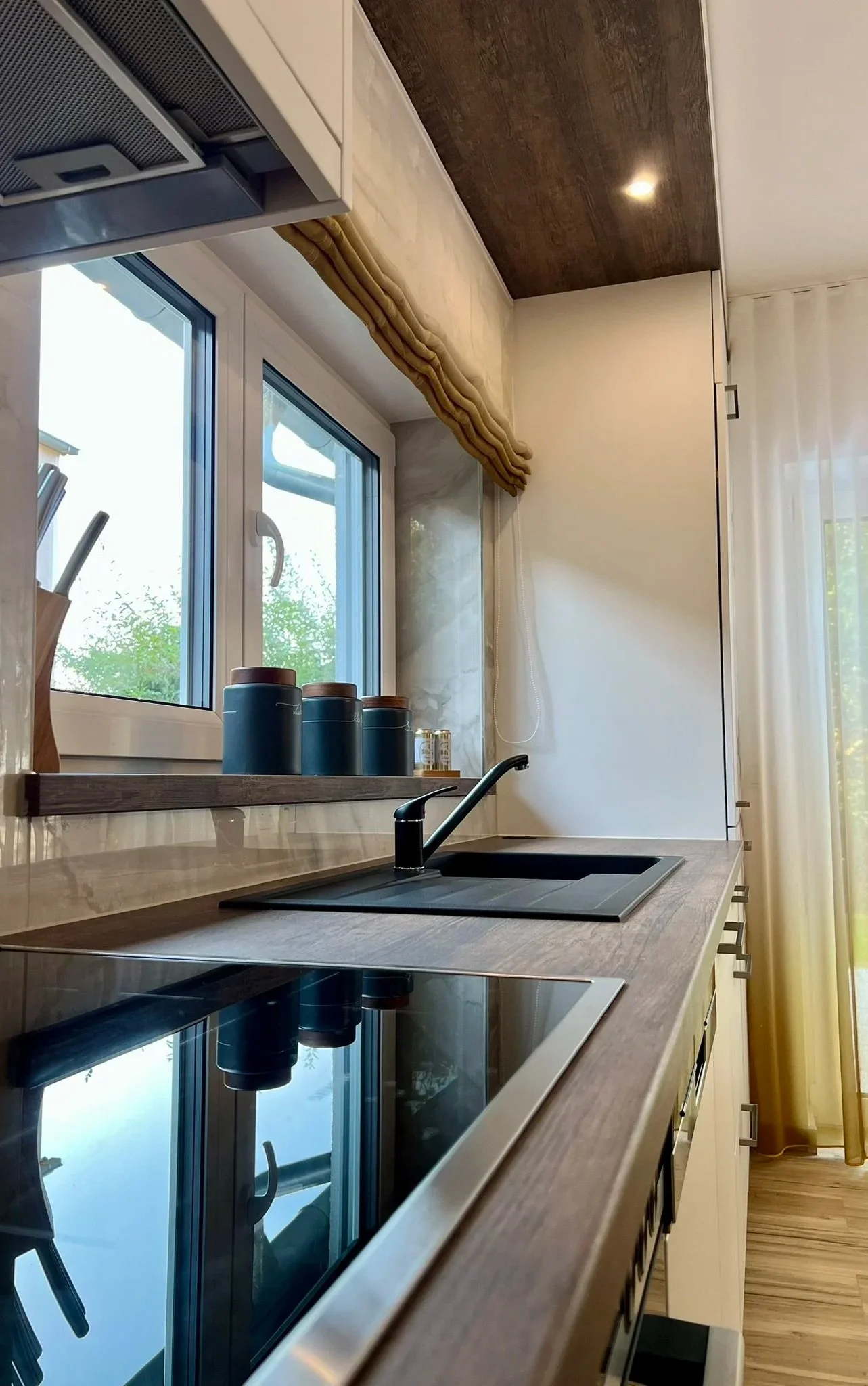 Moderne Küchenzeile mit Holzarbeitsplatte, Herd, Wasserhahn und Fenster mit Blick auf Bäume, beige Vorhänge und helle Wand