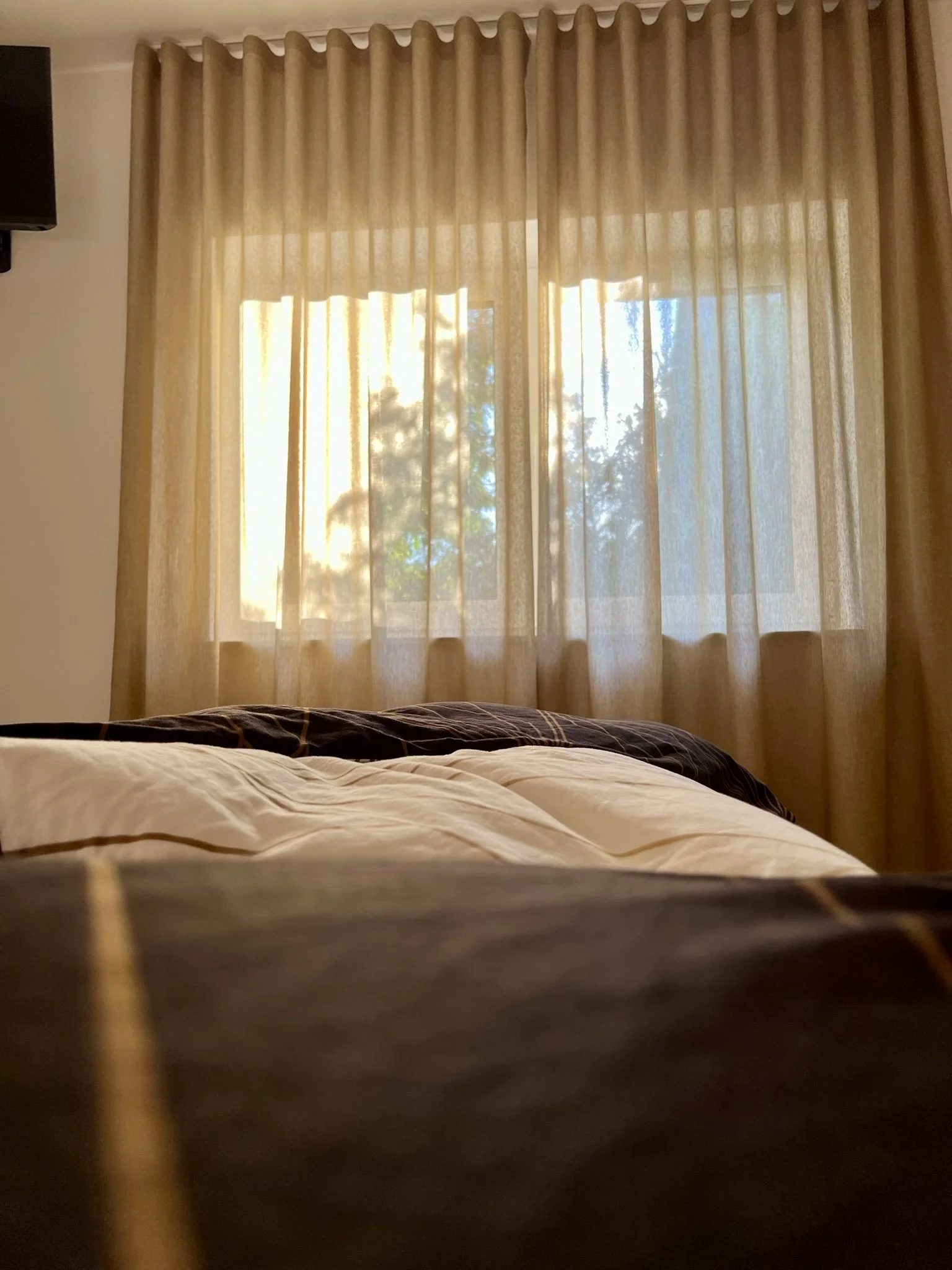 Blick durch ein Fenster mit hellen, beige Vorhängen, Sonnenlicht scheint hindurch, ein Bett im Vordergrund.