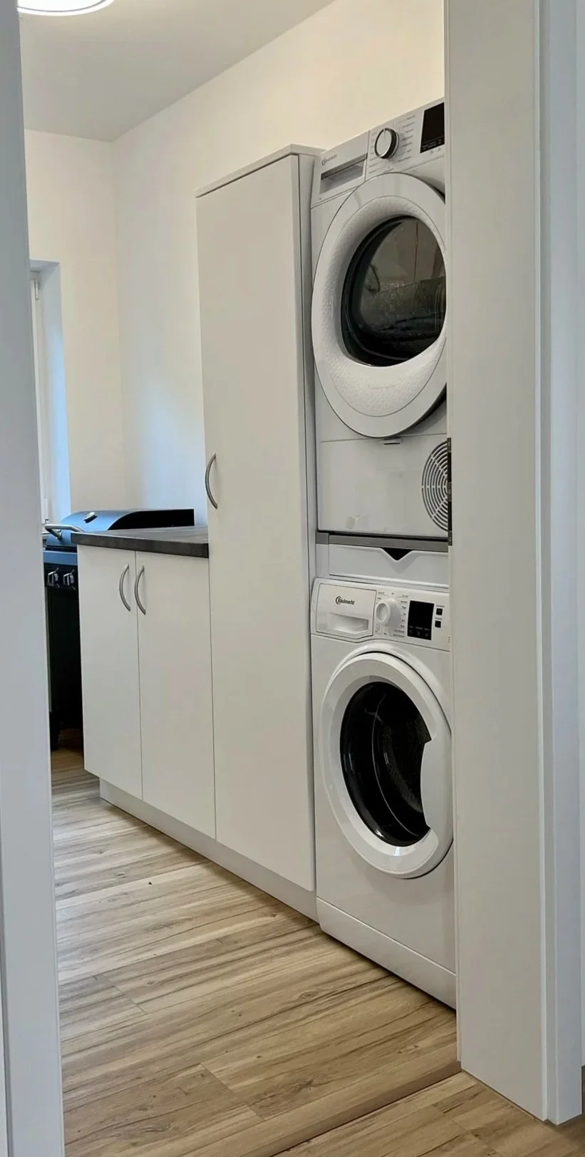 Waschmaschine und Wäschetrockner übereinander in einem kleinen Raum, der durch eine Tür sichtbar ist. Im Hintergrund ein Teil der Küche mit weißen Schränken und Holzfußboden.