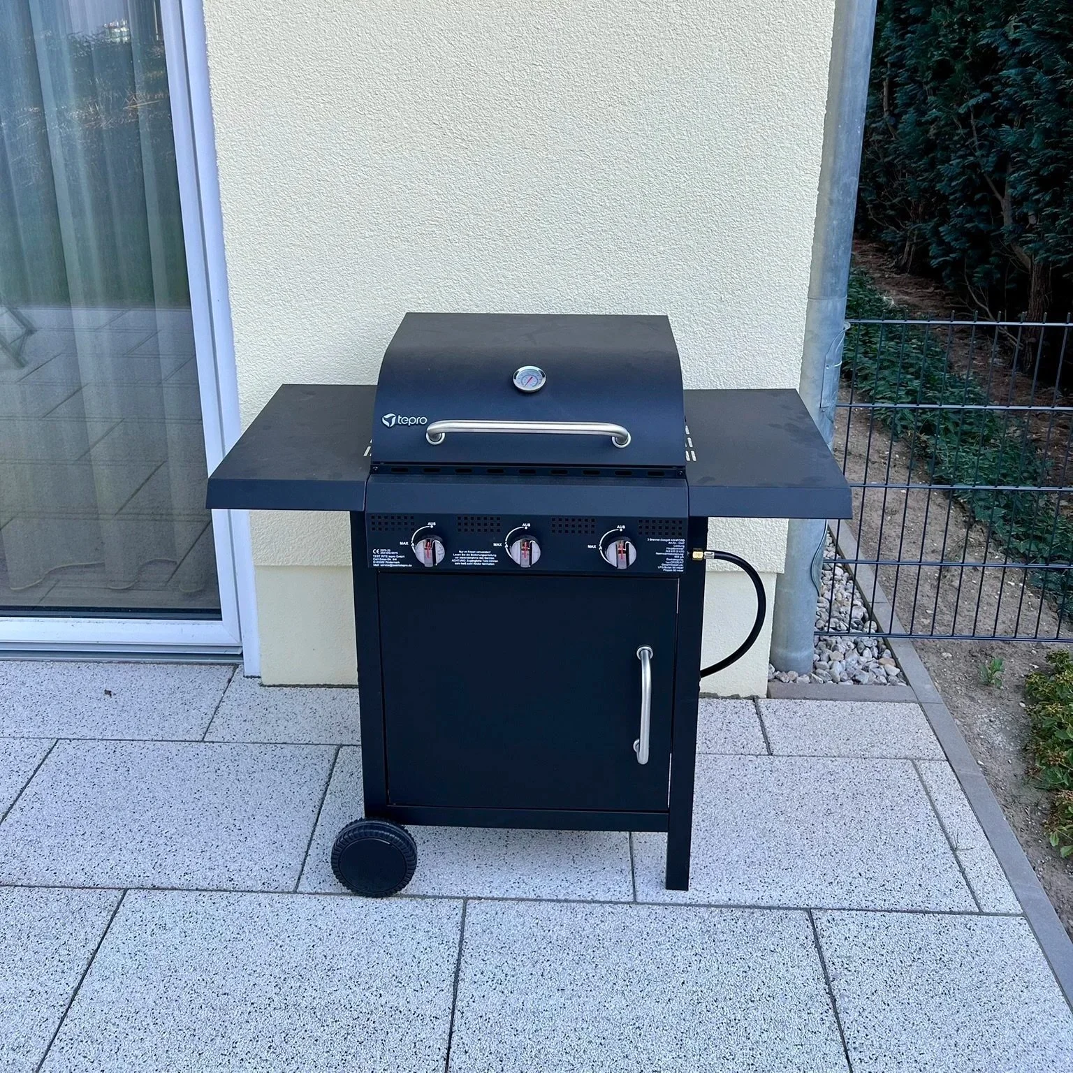 Ein schwarzer Gasgrill mit Drehknöpfen, einem Thermometer und Seitentischen, steht auf einer Terrasse vor einer Hauswand und einem Gitterzaun.