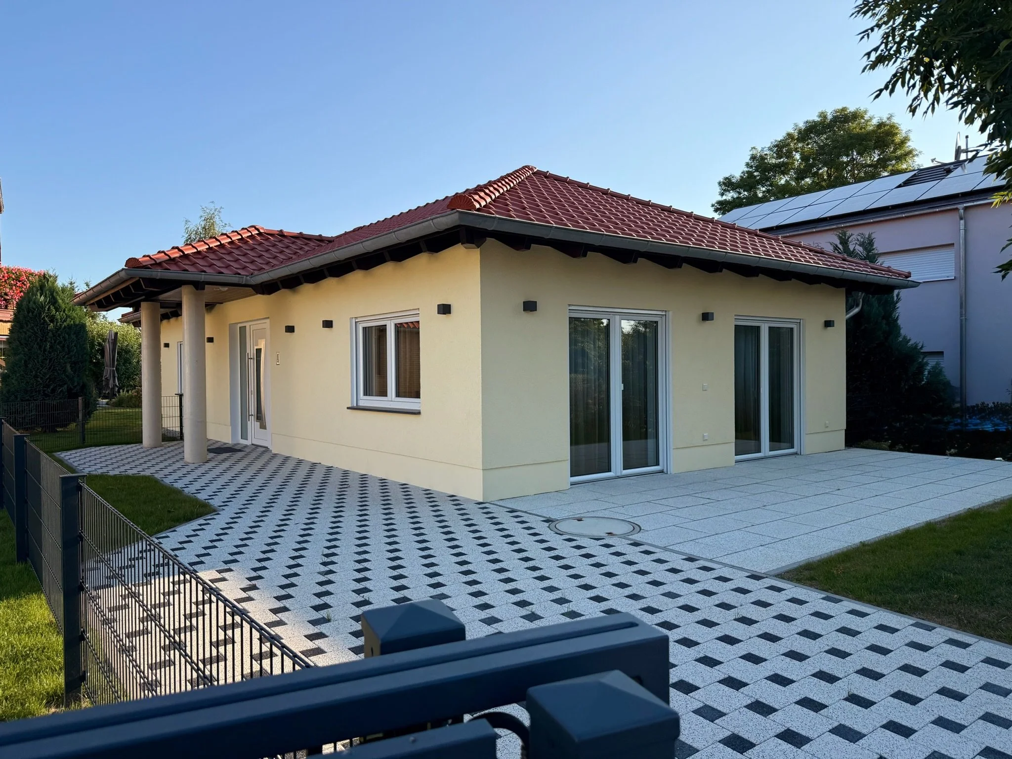 Häuschen mit rotem Ziegeldach, großen Fenstern und einer gepflterten Terrasse, umgeben von einem Garten mit Zaun und Bäumen, sonniger Himmel.