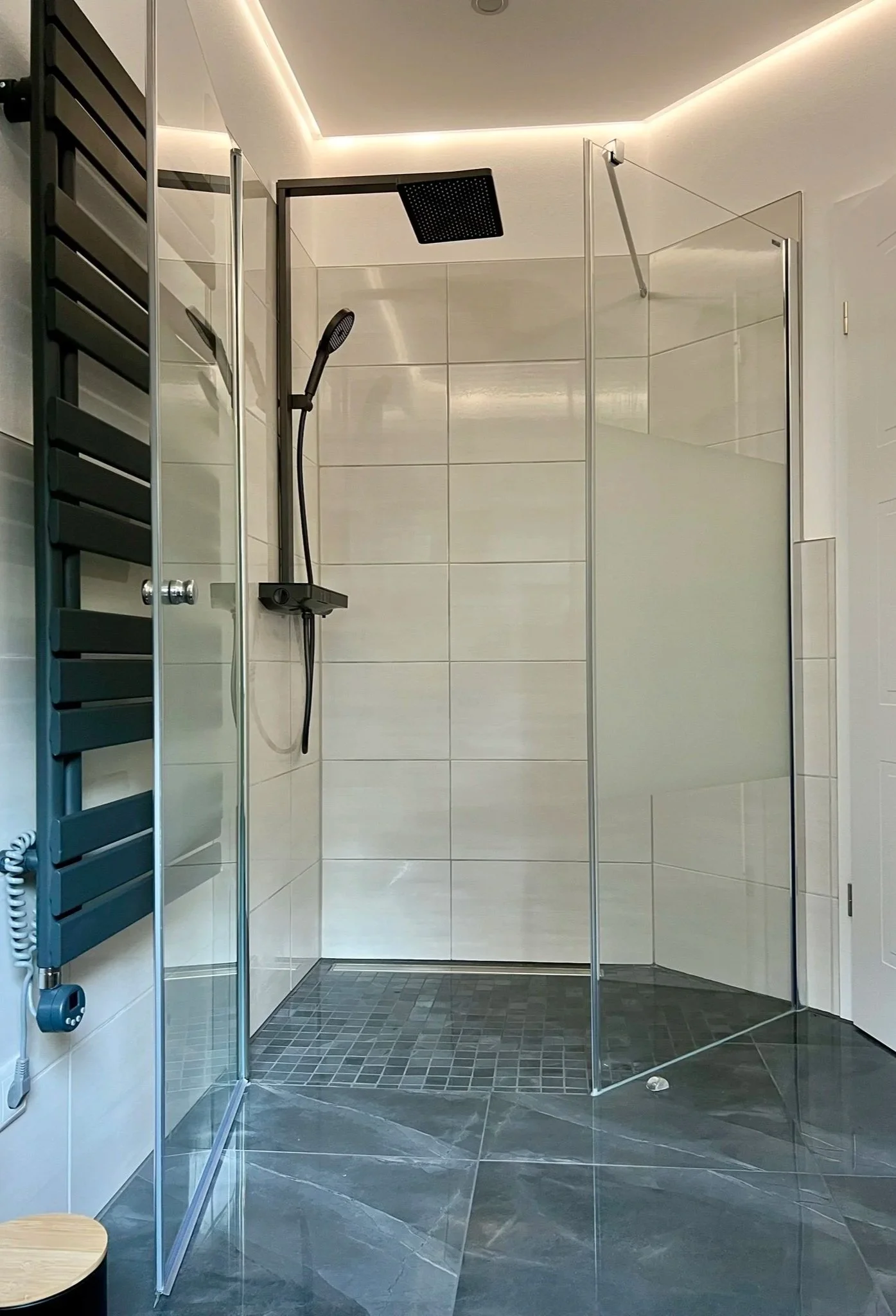 Moderne, offene Duschkabine mit dunklem Boden und großem, quadratischem Duschkopf an der Decke, umgeben von weißen Wandfliesen und einer Glastür.