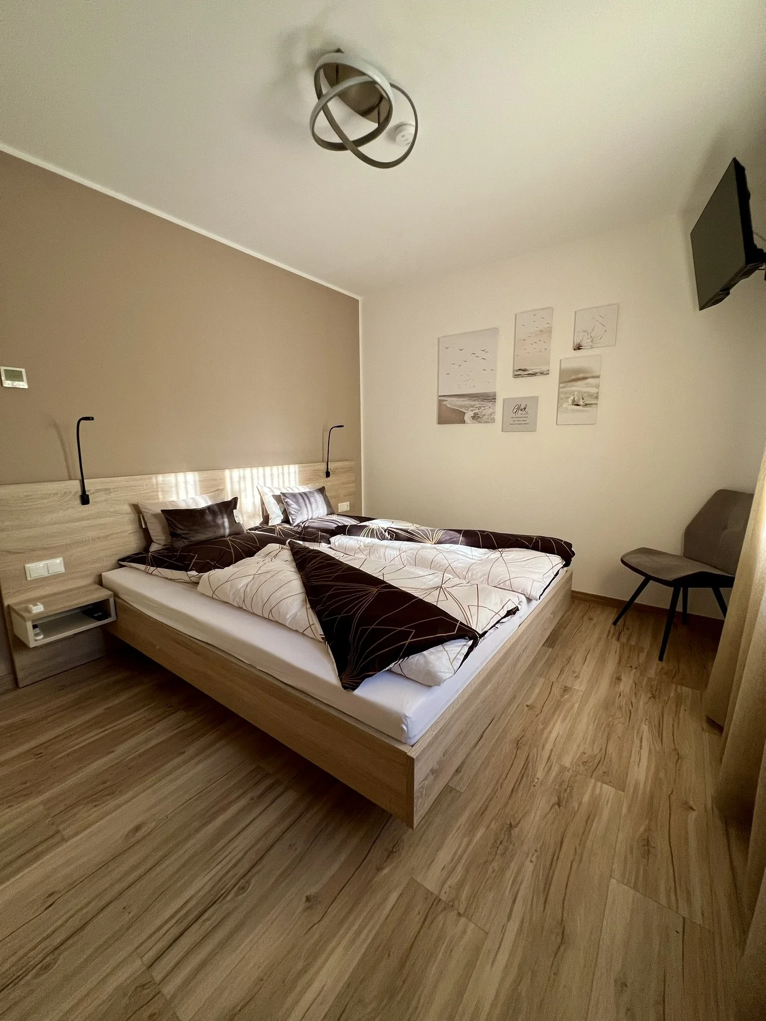 Ein Schlafzimmer mit Doppelbett, Nachttischen, Wandlampen, Wanddeko, Sessel, Fernseher und Holzboden.