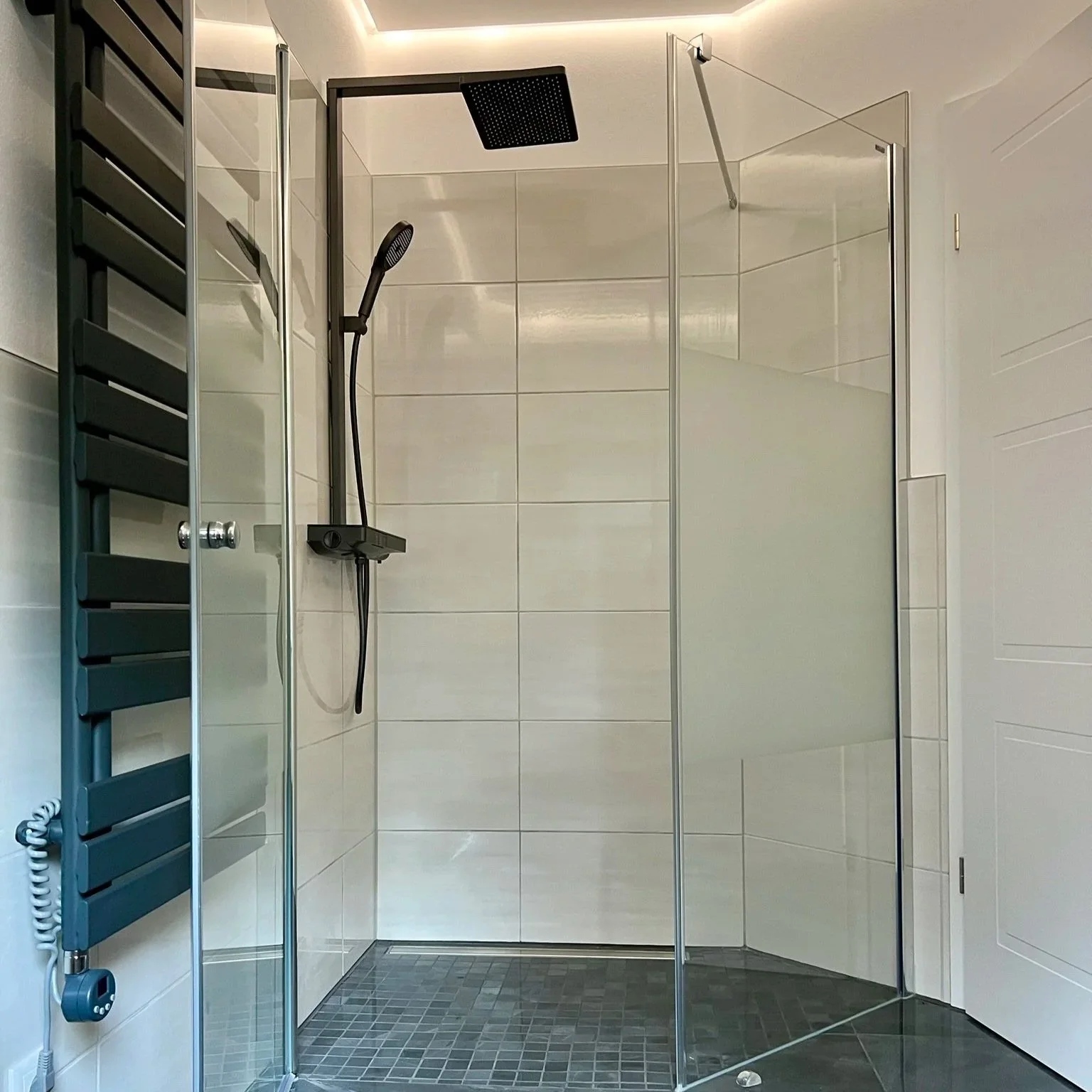 Moderner Badezimmer-Duschbereich mit begehbarer Dusche, schwarzem Duschkopf, Handbrause, gläserner Tür, weißen Wandfliesen und dunklem Boden, daneben Heizkörper.