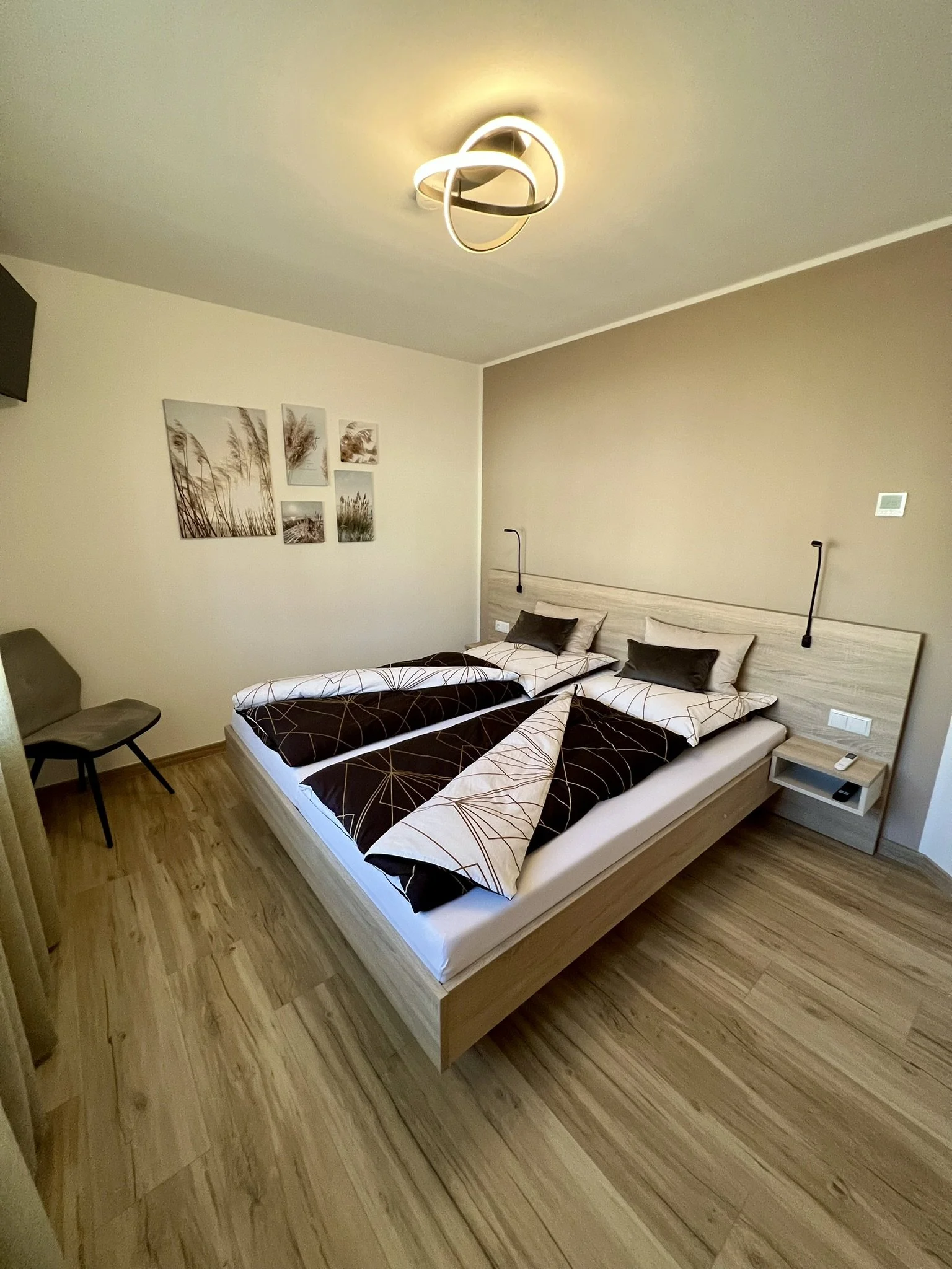 Ein Schlafzimmer mit einem Doppelbett, das mit schwarz-weiß gemusterten Decken und Kissen bedeckt ist. Es gibt eine Nachttischlampe, ein Nachttisch mit einem Mobiltelefon und eine Wanddekoration aus fünf Bilderrahmen mit Naturmotiven. Die Zimmerdecke hat eine moderne Leuchte.