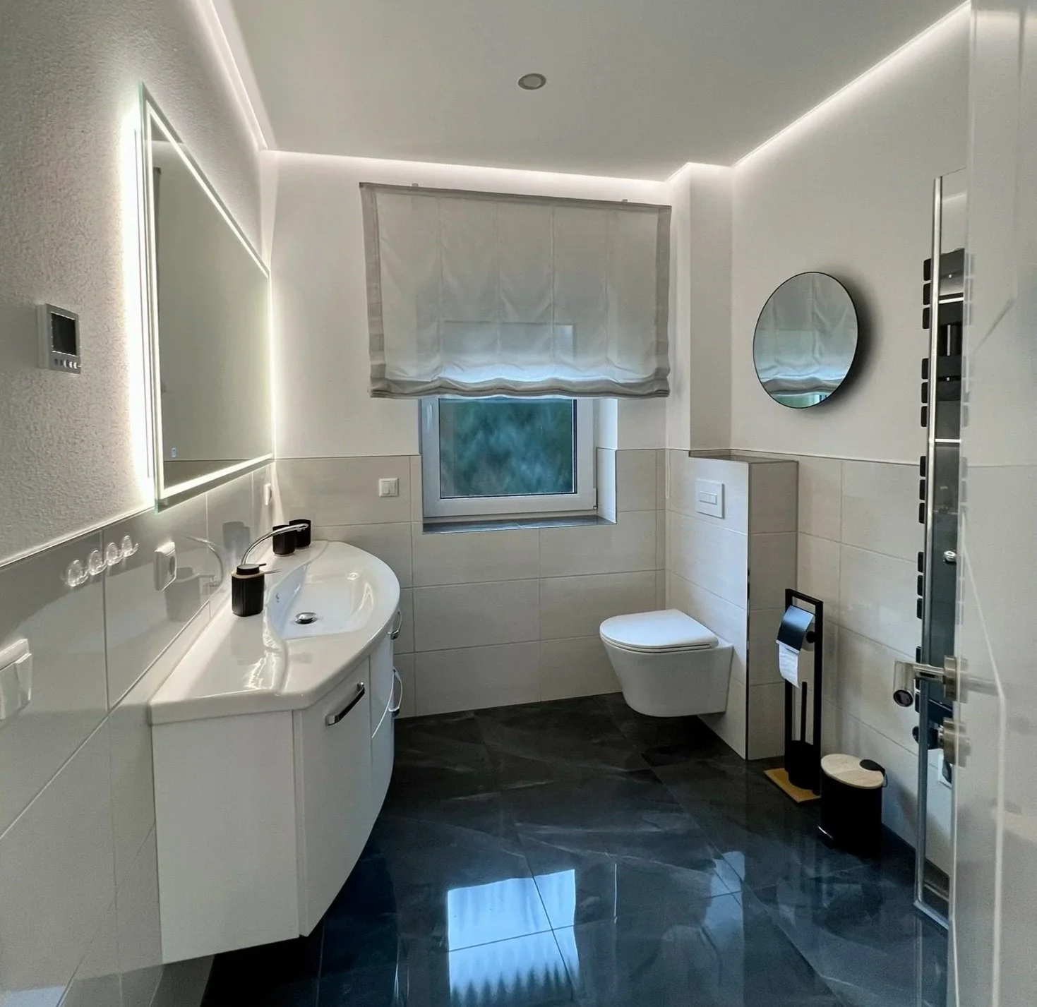 Modernes Badezimmer mit Waschbecken, Spiegel, Toilette, Fenster mit Vorhang, dunklem Bodenfliesen, und Dusche mit Glasfront.