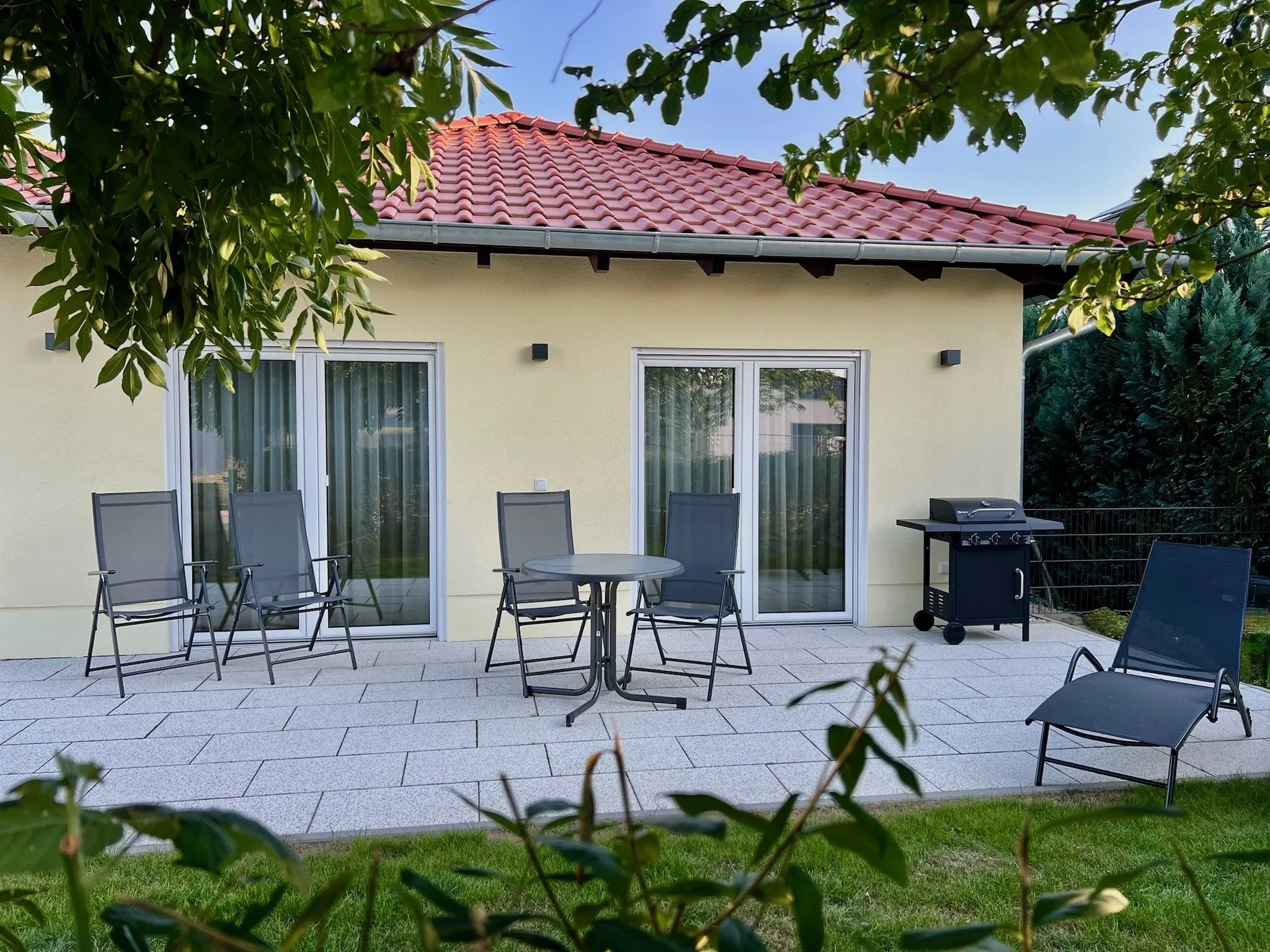Auf einer Terrasse befindet sich ein runder Tisch mit Stühlen, im Hintergrund eine gelbe Hauswand mit zwei großen Fenstern und einer Haustür, ein Grill steht an der Wand, umgeben von Bäumen und Sonnenschein.