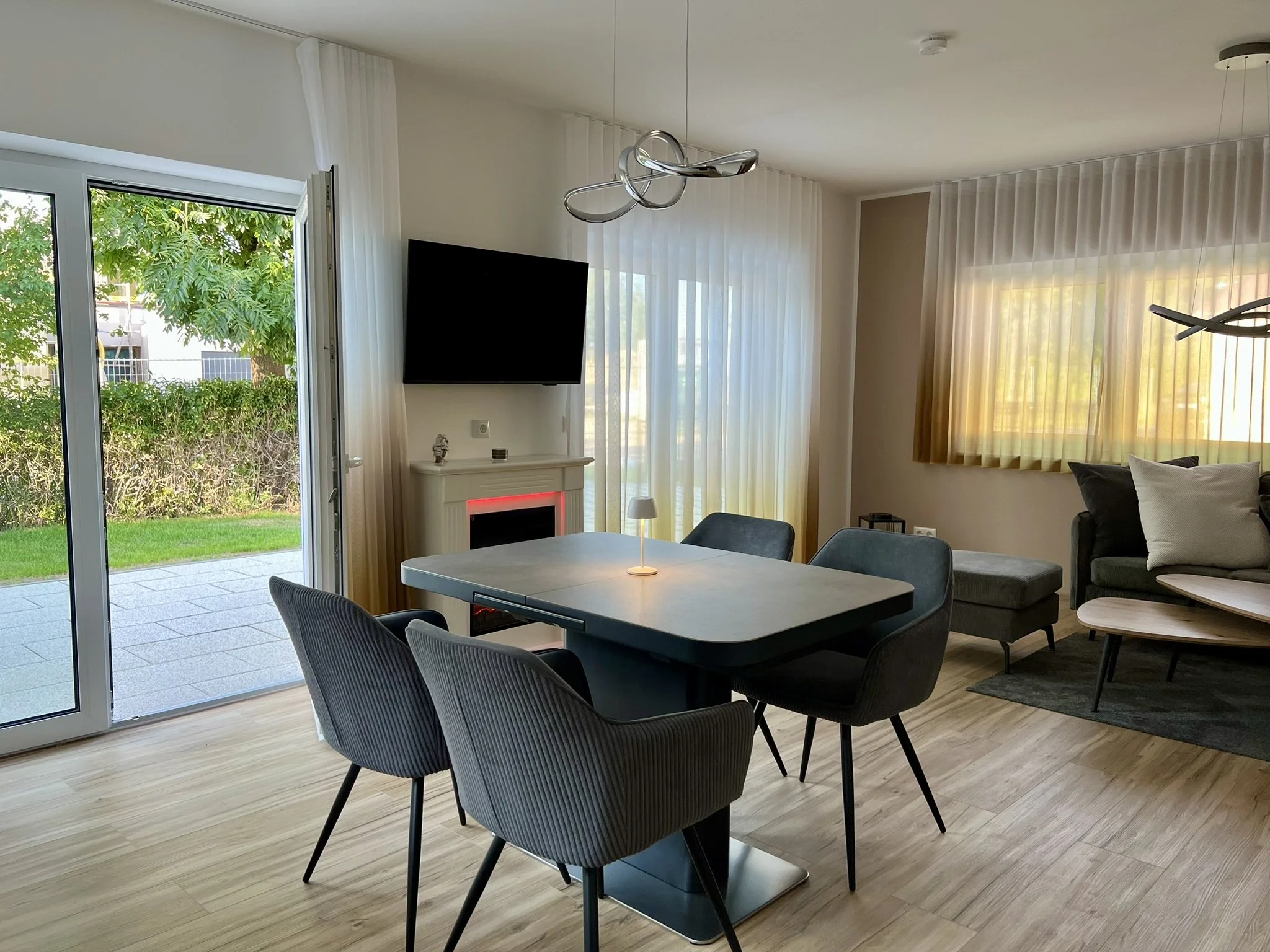 Moderner Wohnzimmerbereich mit Esstisch, fünf schwarze gepolsterte Stühle, große Fenster mit weißen Vorhängen, Fernseher, Kamin mit rotem Licht, dekorare Figuren, Couch mit Kissen, beige Wand, Holzfußboden.