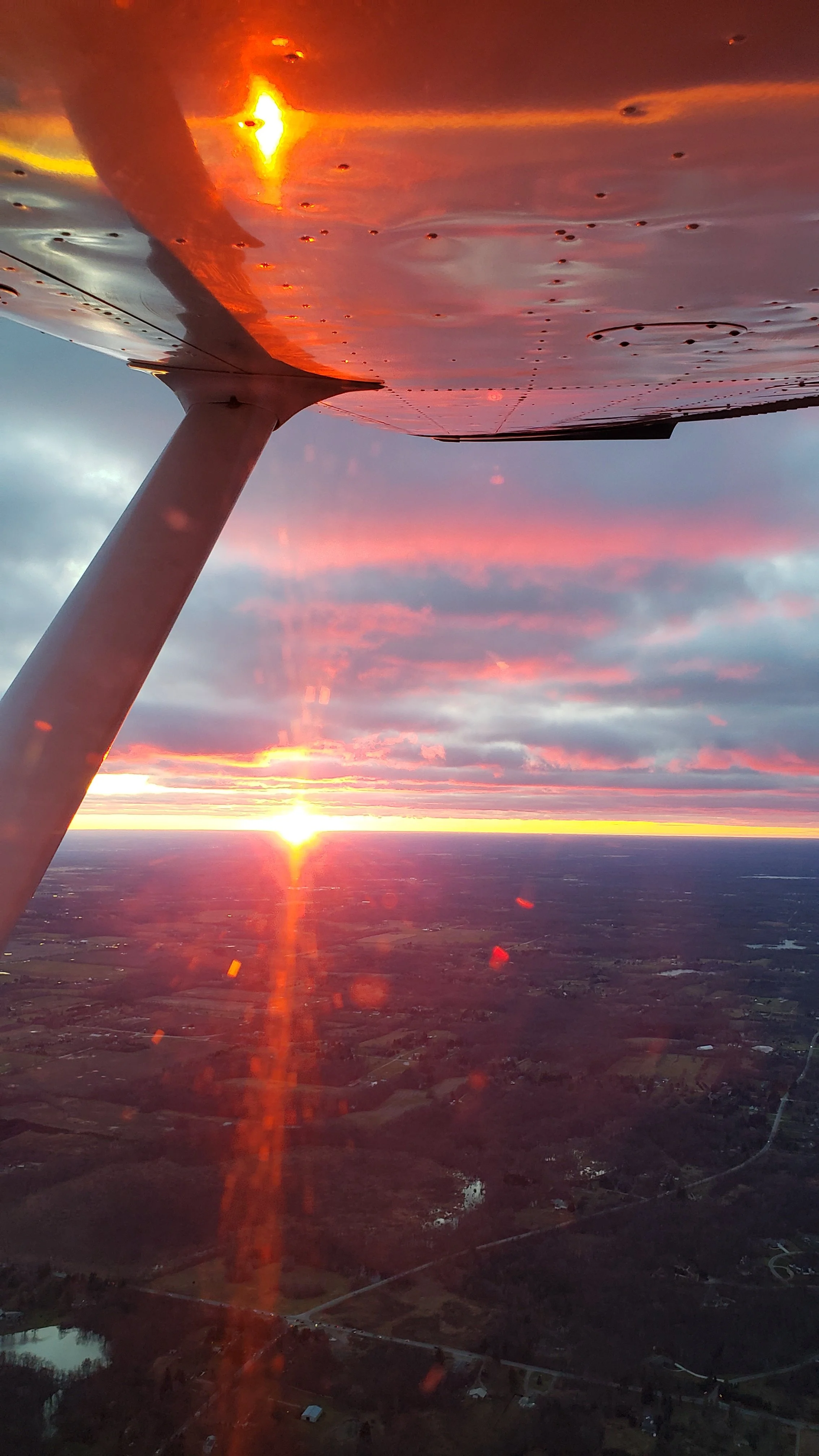 Sunset viewed from the wing of an airplane, with the aircraft's wing and support strut visible in the foreground. The sky is filled with colorful clouds, casting a warm glow over the landscape below.